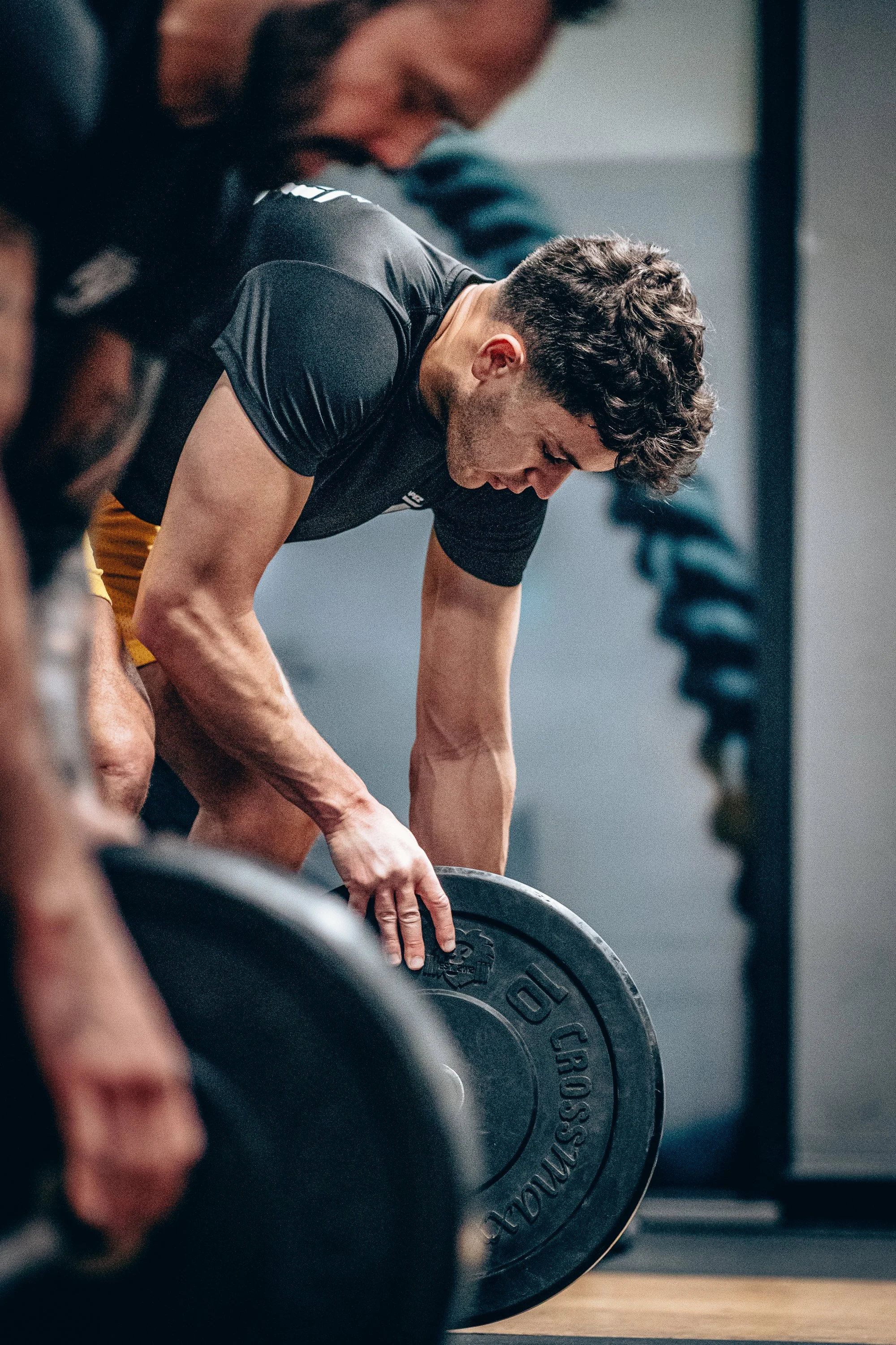 A man performing a deadlift exercise in a gym, lifting a barbell with weights. He is focused, bending forward with his hands gripping the barbell, wearing a black shirt and yellow shorts.