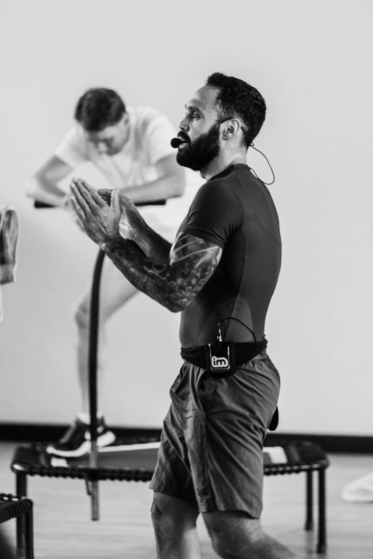 Man met een microfoon en een smartwatch die oefeningen doet in een fitnessruimte, met een persoon op een trampoline op de achtergrond.