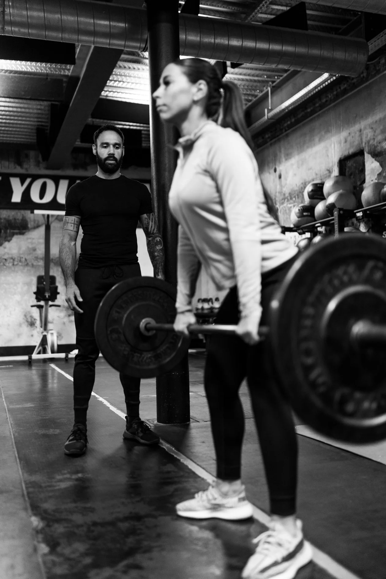 Een vrouw tiltert een haltergewicht in een sportschool, terwijl een man haar coacht of observeert, met gewichten en fitnessapparatuur op de achtergrond.