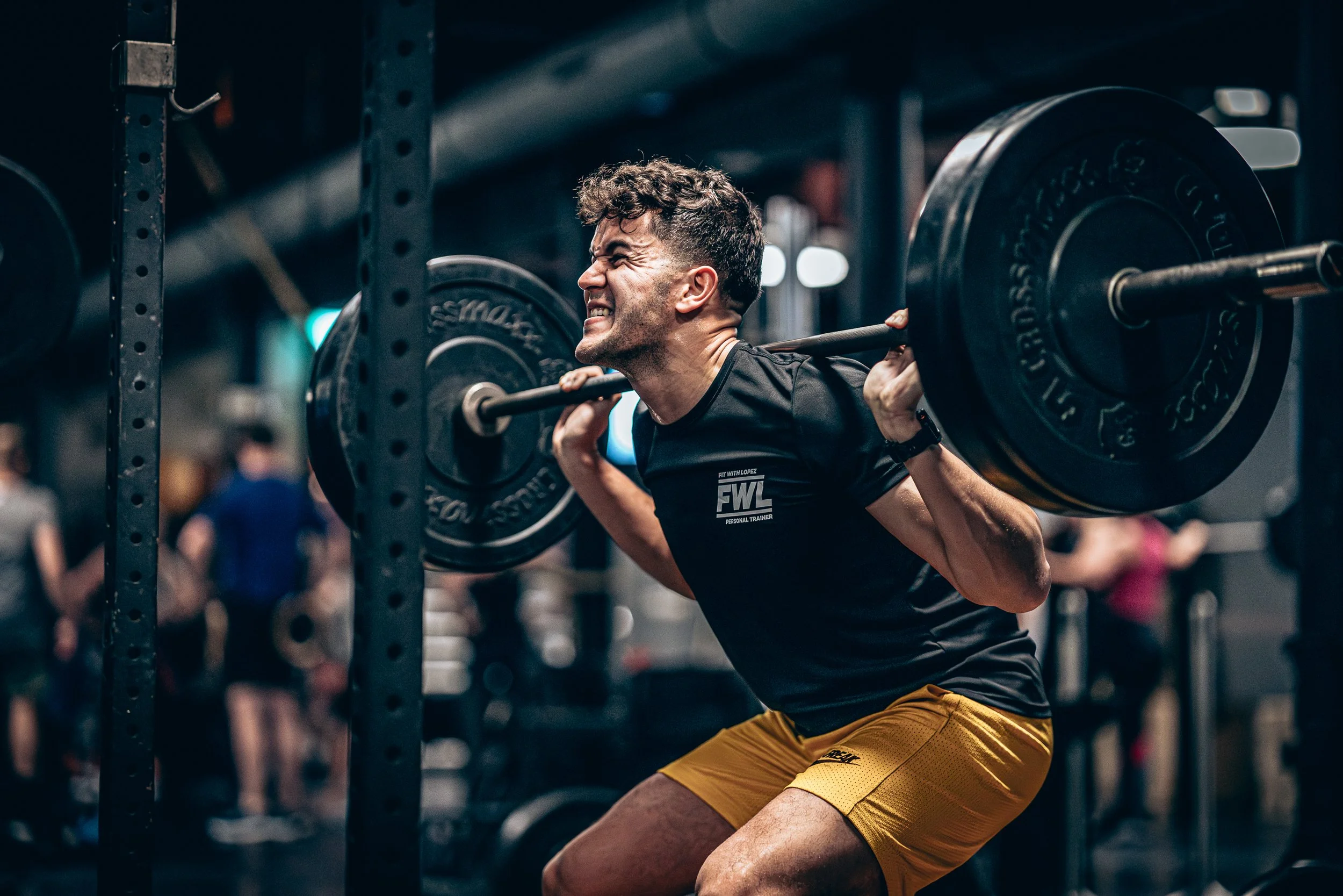 A man lifting a barbell in a gym with a strained expression on his face, wearing a black t-shirt and yellow shorts.