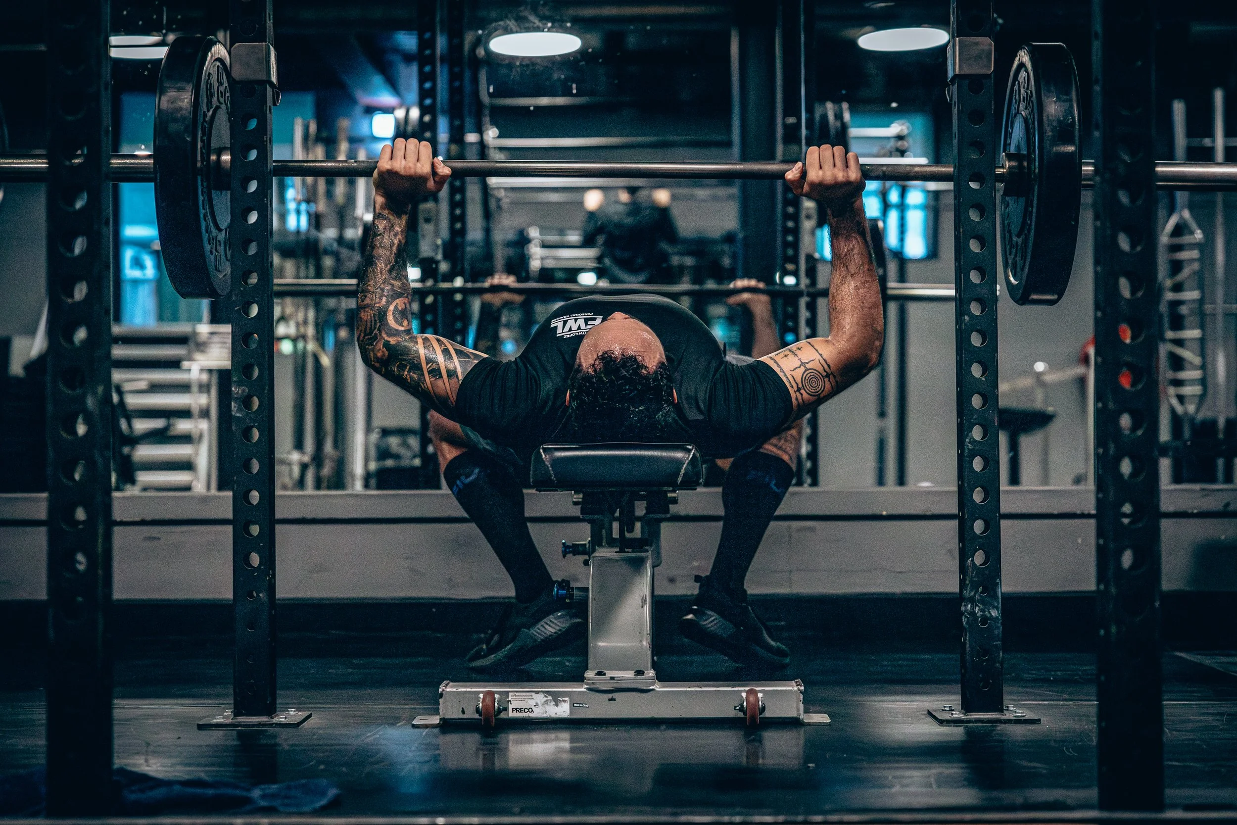 A man with tattoos is lying on a bench, lifting a barbell with weights in a gym.