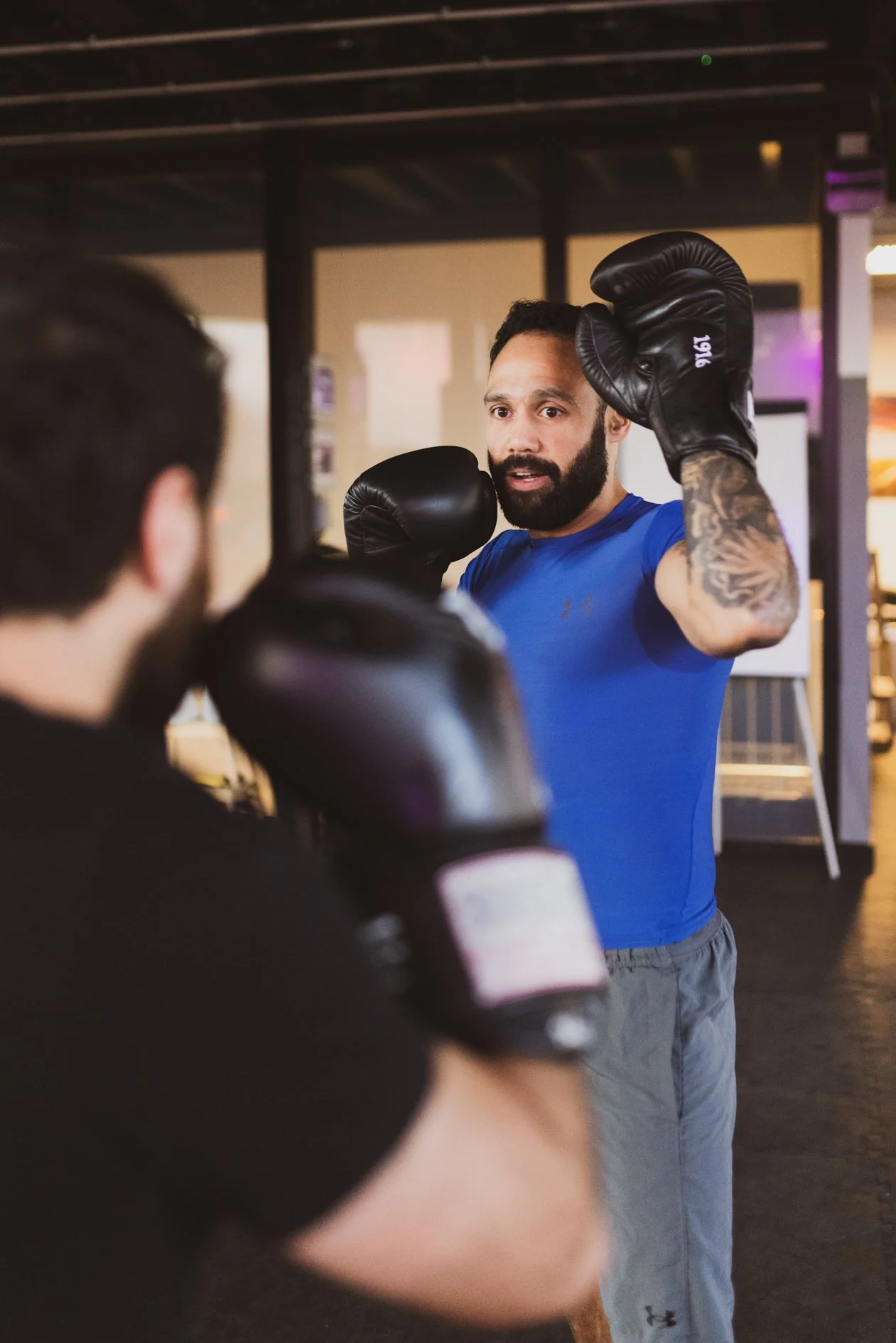 Een man met een baard en een blauw sportshirt leert iemand anders boksen met behulp van beschermende handschoenen in een sportschool.