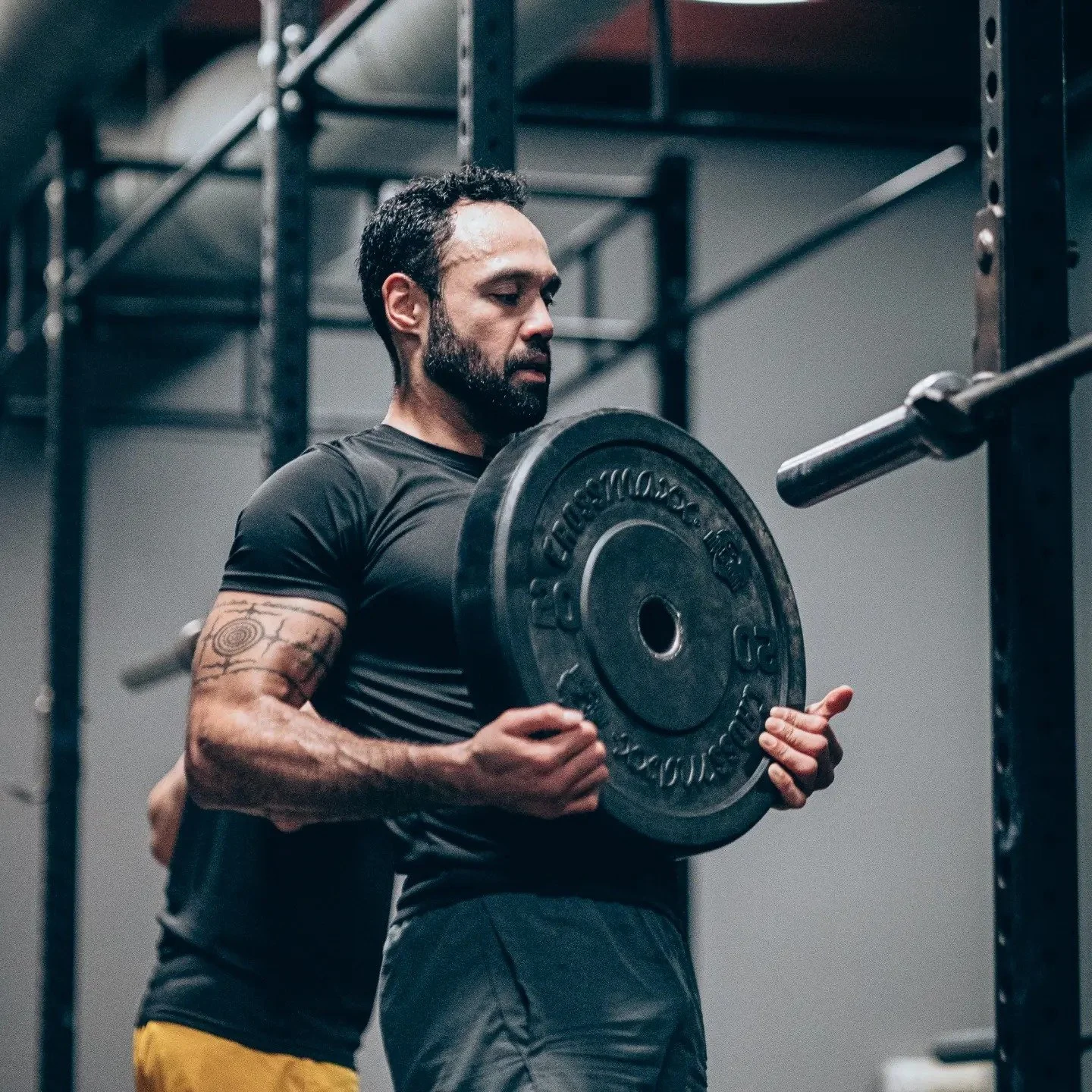 A man with a beard and tattoos on his arm lifting a weight plate in a gym.