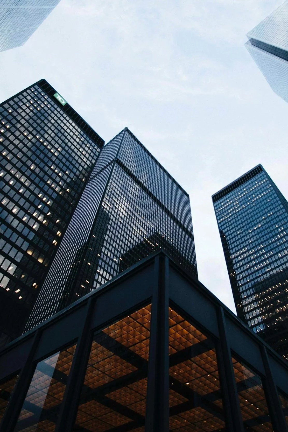 Looking up at several tall glass skyscrapers in a city skyline, with a glass-roofed structure in the foreground.