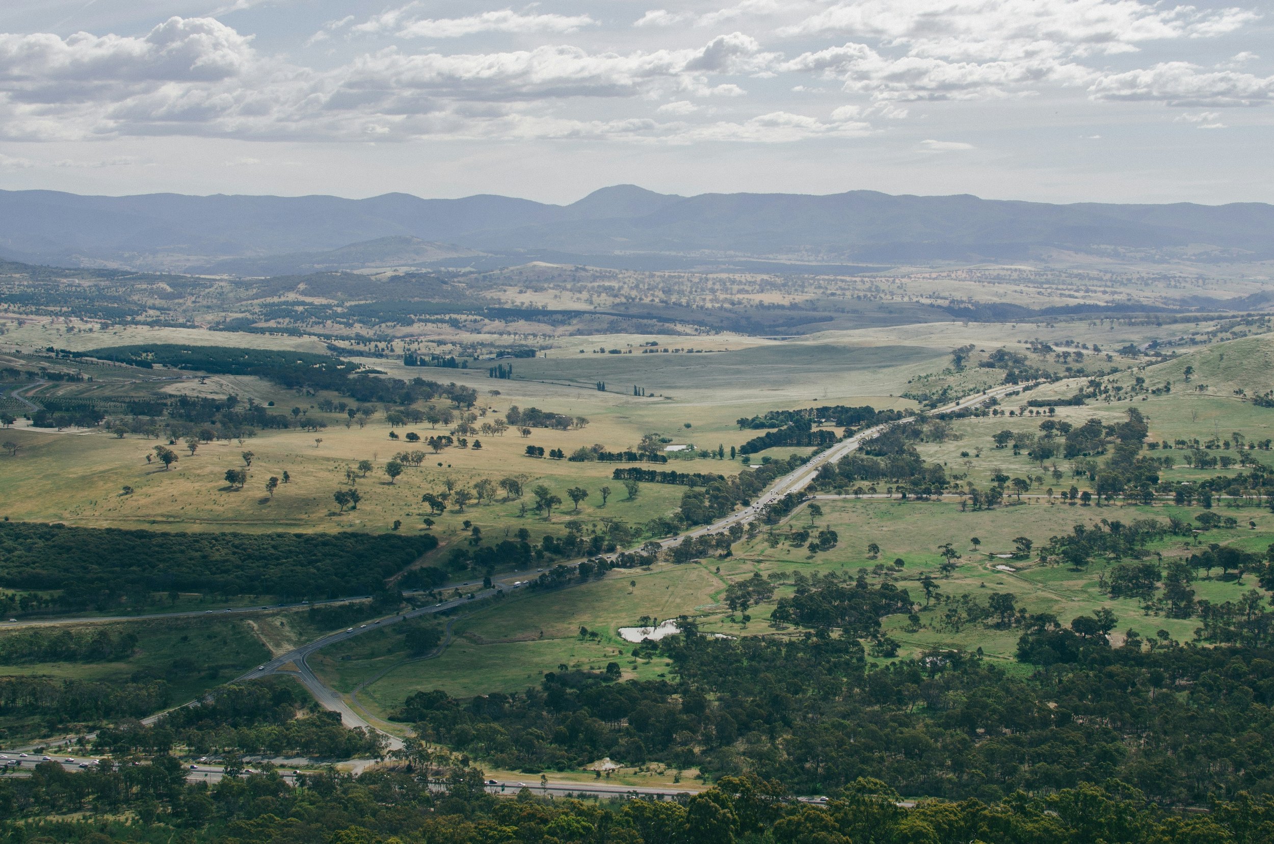 A vast landscape of rolling green hills with patches of trees, roads, and a distant mountain range under a partly cloudy sky.