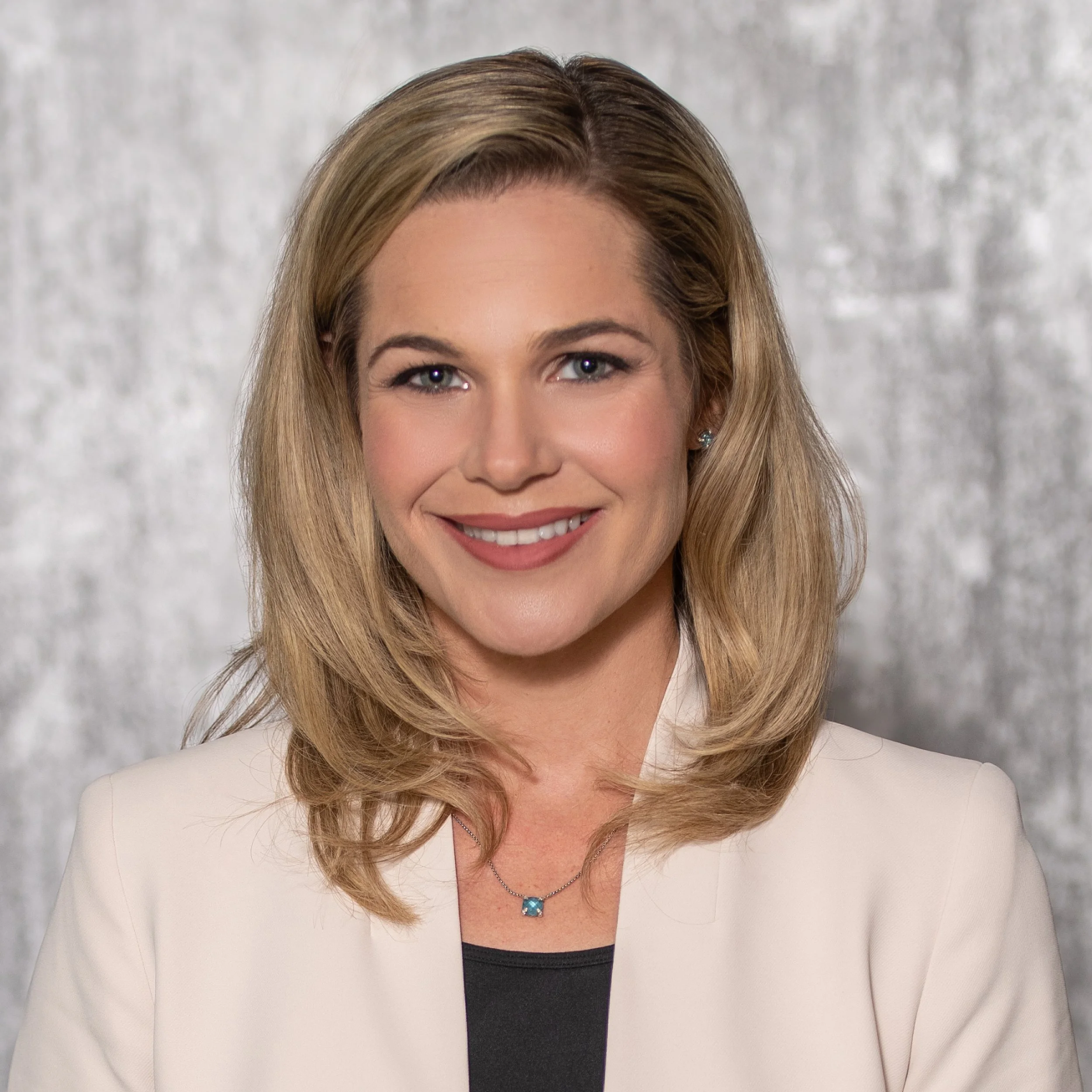 Professional woman with blonde hair smiling, wearing a white blazer and a necklace, with a silver background