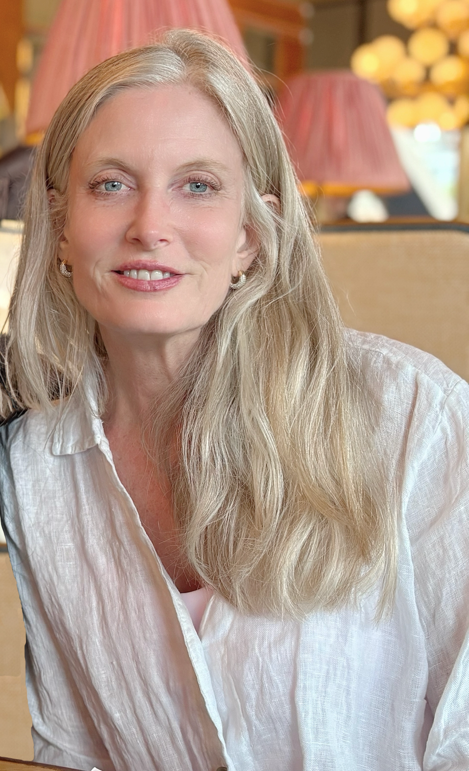 A woman with long blonde hair and blue eyes smiling, sitting in a cozy indoor setting with warm lighting and pink lampshades in the background.