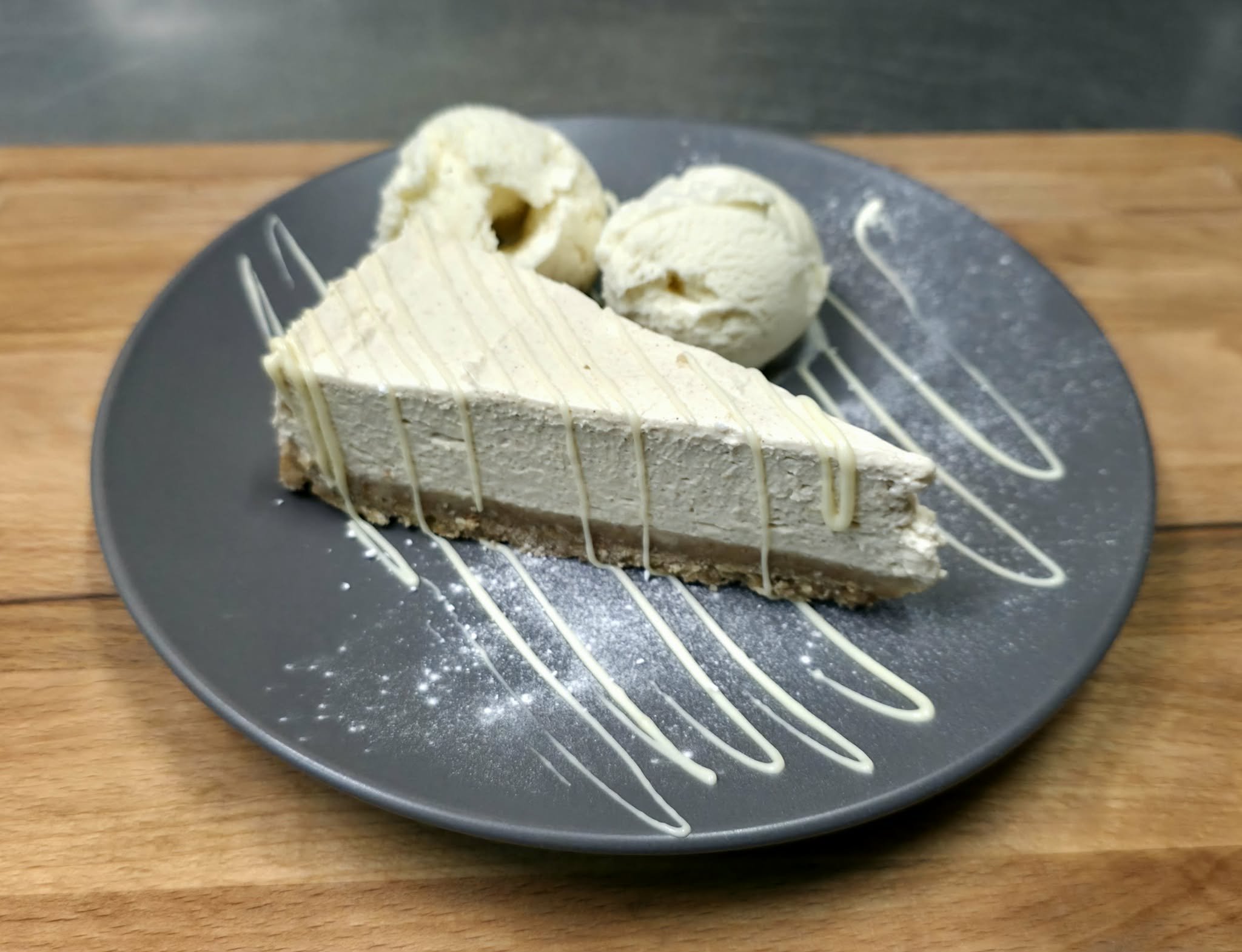 Slice of cheesecake with two scoops of vanilla ice cream on a dark plate with white chocolate drizzle and powdered sugar, on a wooden surface.