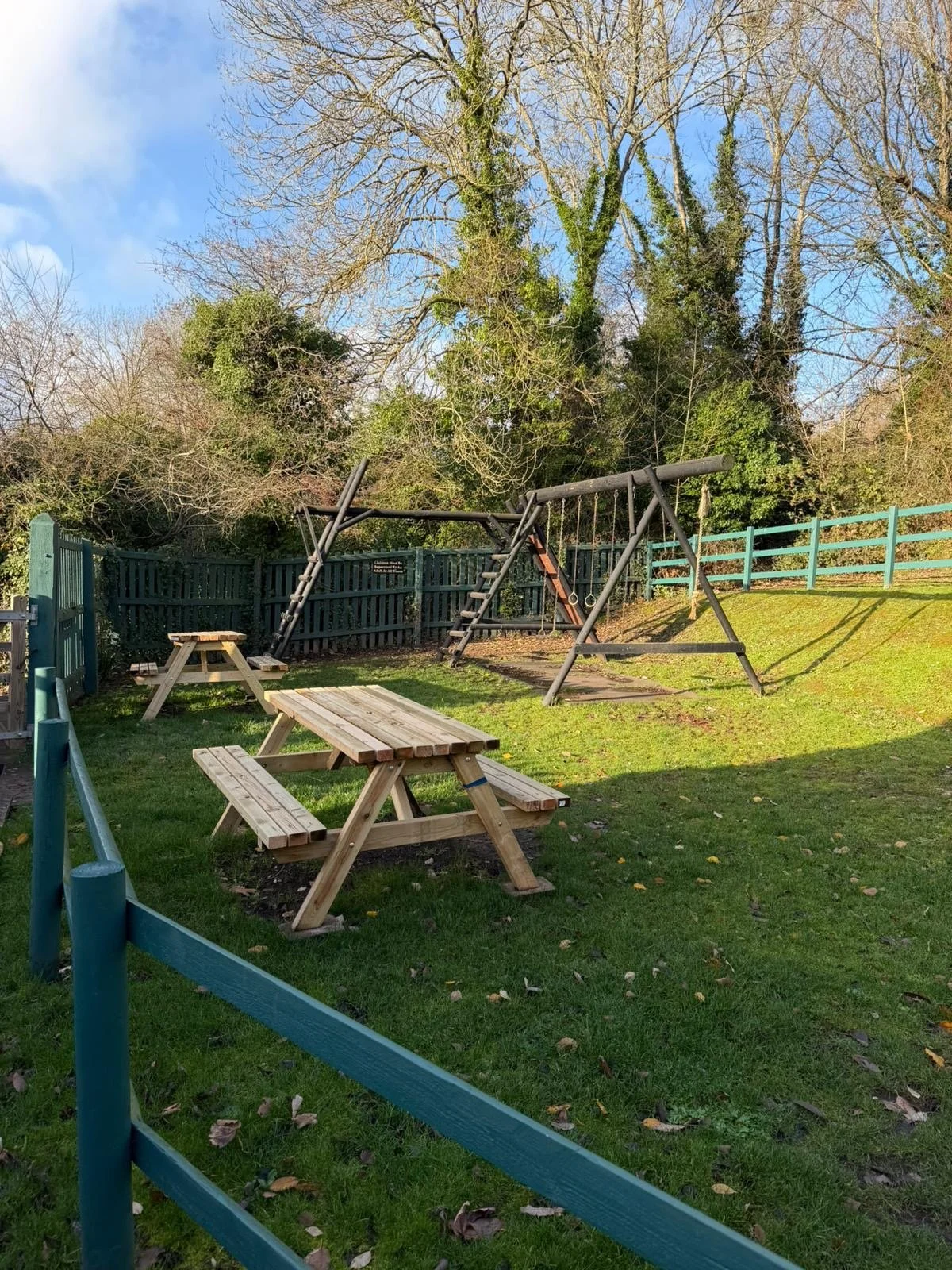 A small playground area with a picnic table, a wooden slide, and swings, enclosed by a green fence with tall trees and a blue sky in the background.