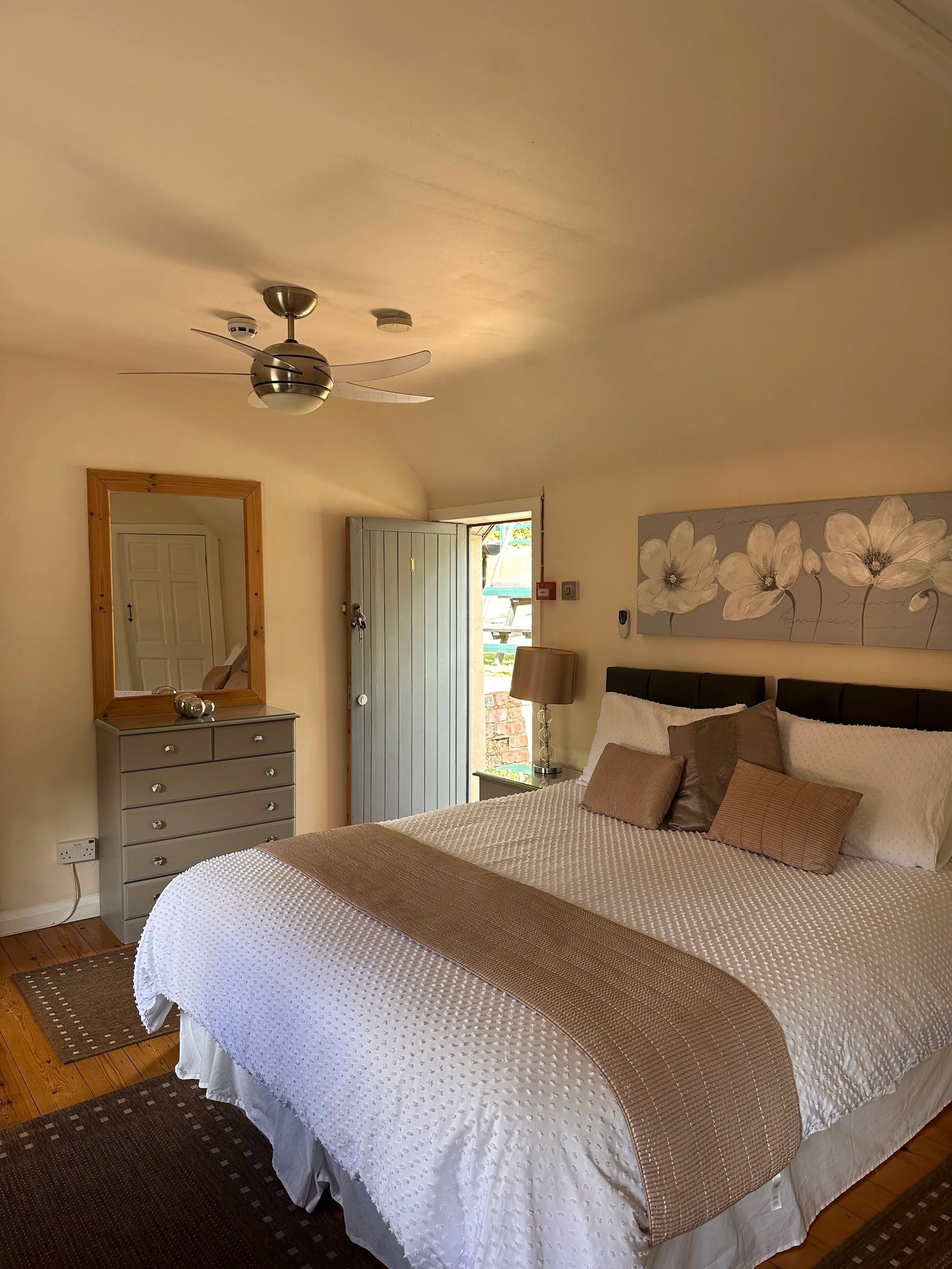 A bedroom with a large bed, multiple pillows, a gray dresser with a mirror, a table lamp, and an open door leading outside. There is a ceiling fan and wall art above the bed.