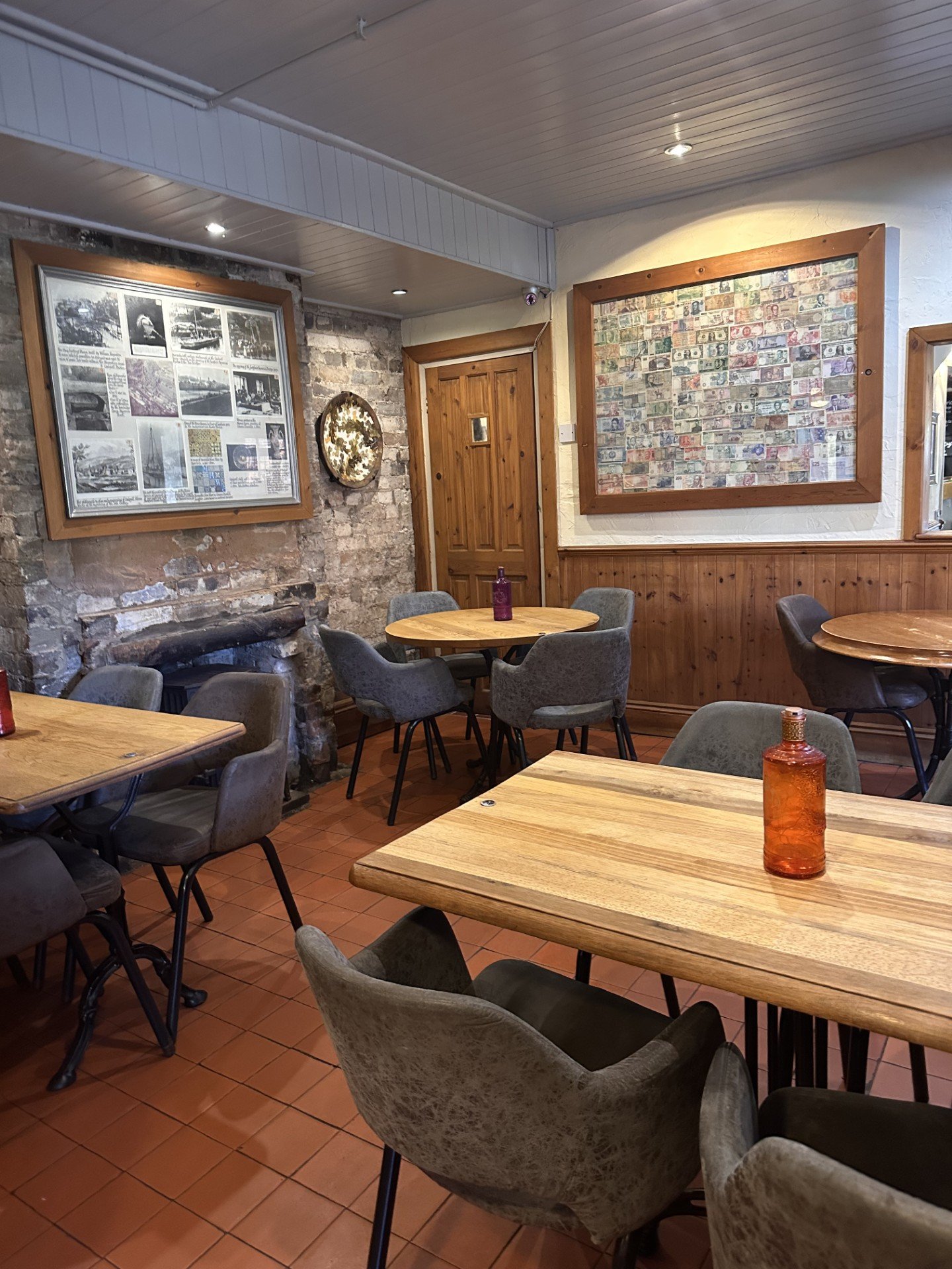 Interior of a cozy restaurant or cafe with wooden tables and gray upholstered chairs, a brick wall with framed historical photos and a display of banknotes, and terracotta floor tiles.