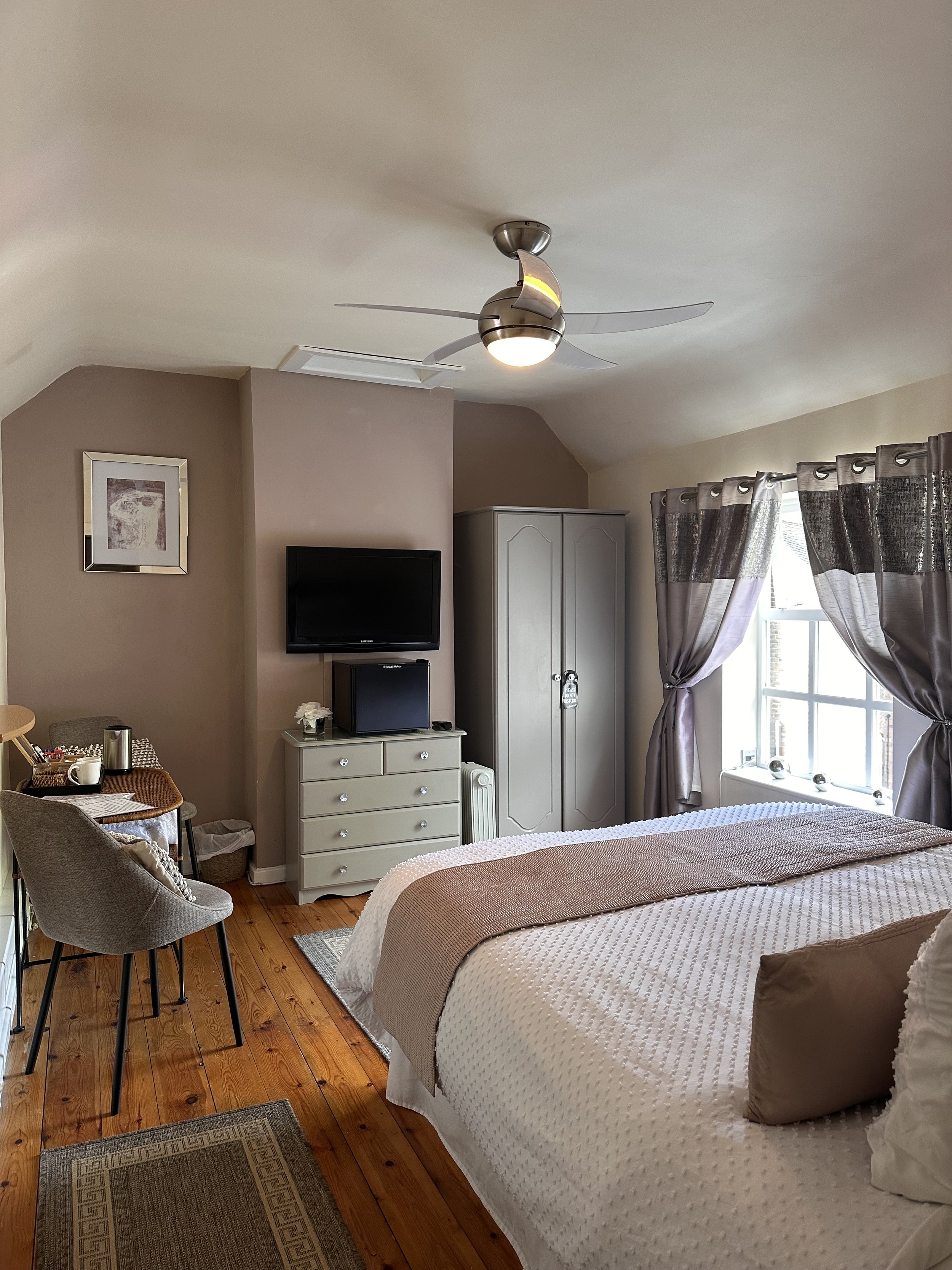 A cozy bedroom with a bed, dresser, wardrobe, and wall-mounted TV. The room features a ceiling fan with lights, a window with curtains, hardwood flooring, and a small sitting area with chairs and a table.