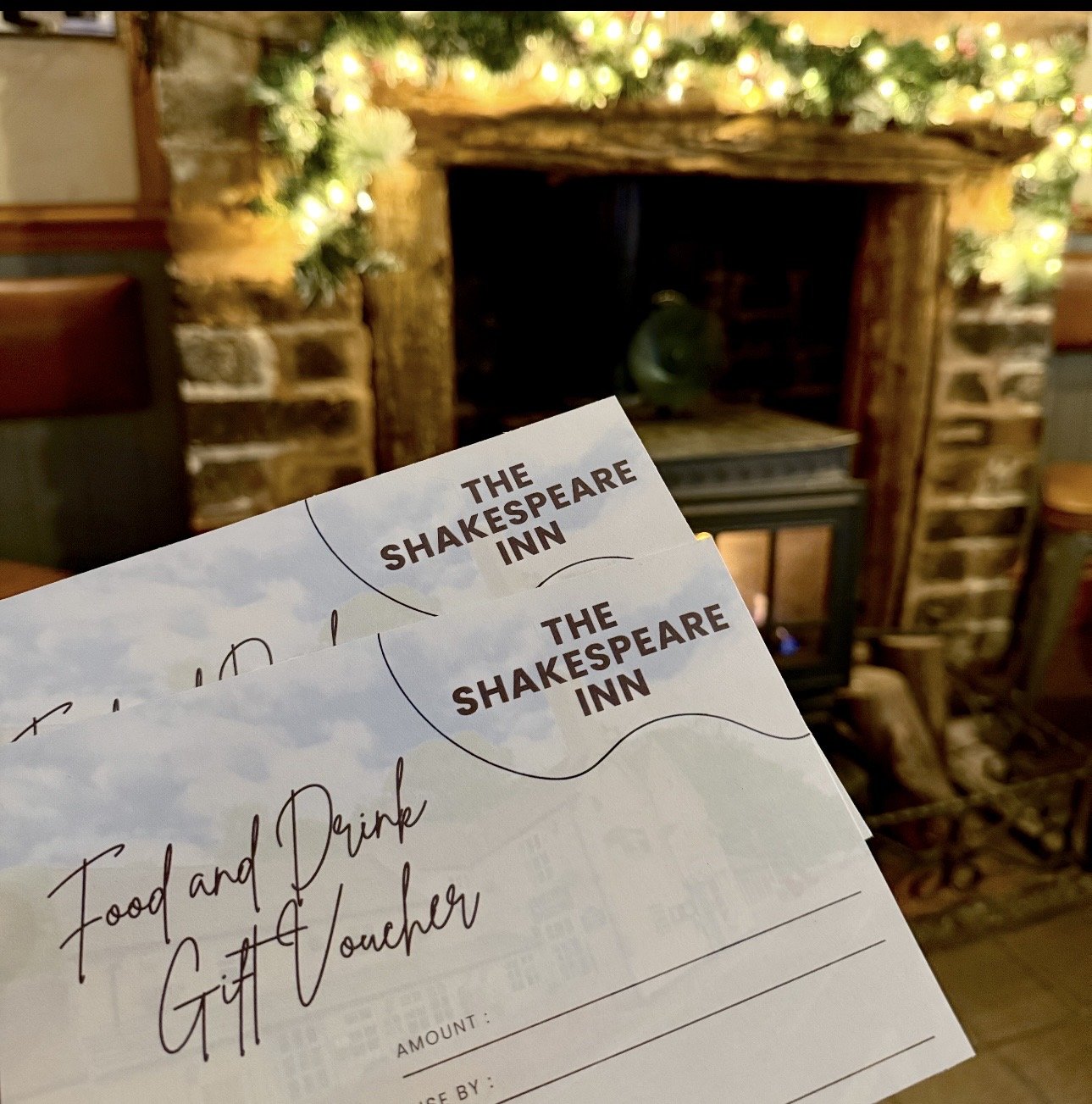 Gift certificates for food and drink from The Shakespeare Inn are held up in front of a decorated fireplace with a mantle adorned with greenery and lights.