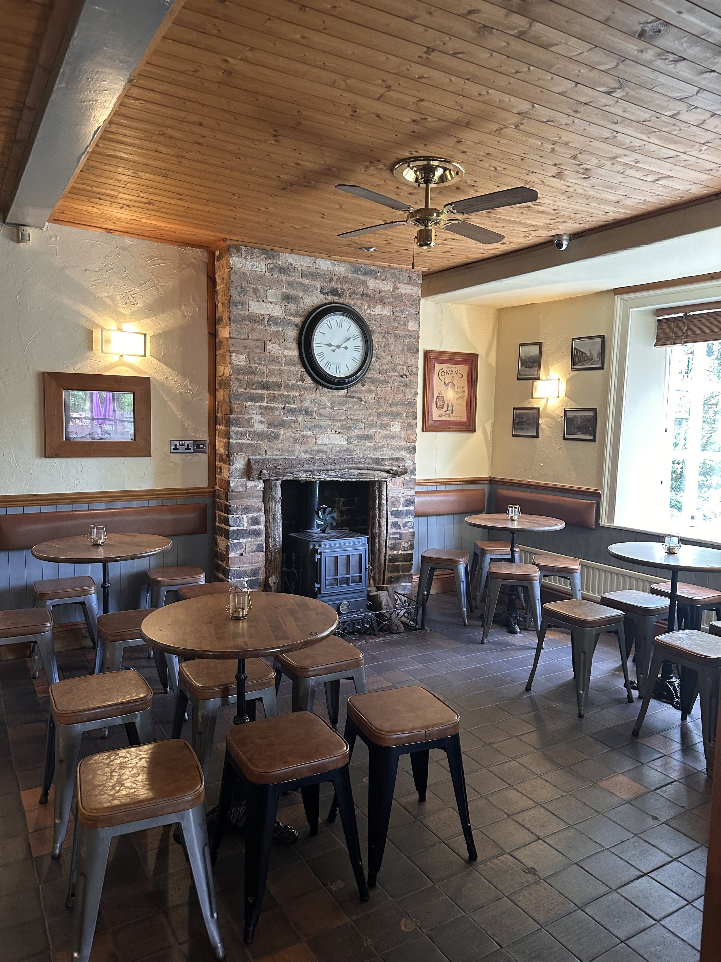 Interior of a cozy pub or cafe with wooden ceiling, brick fireplace, and several round tables with metal and leather stools, decorated with wall art and a large window.