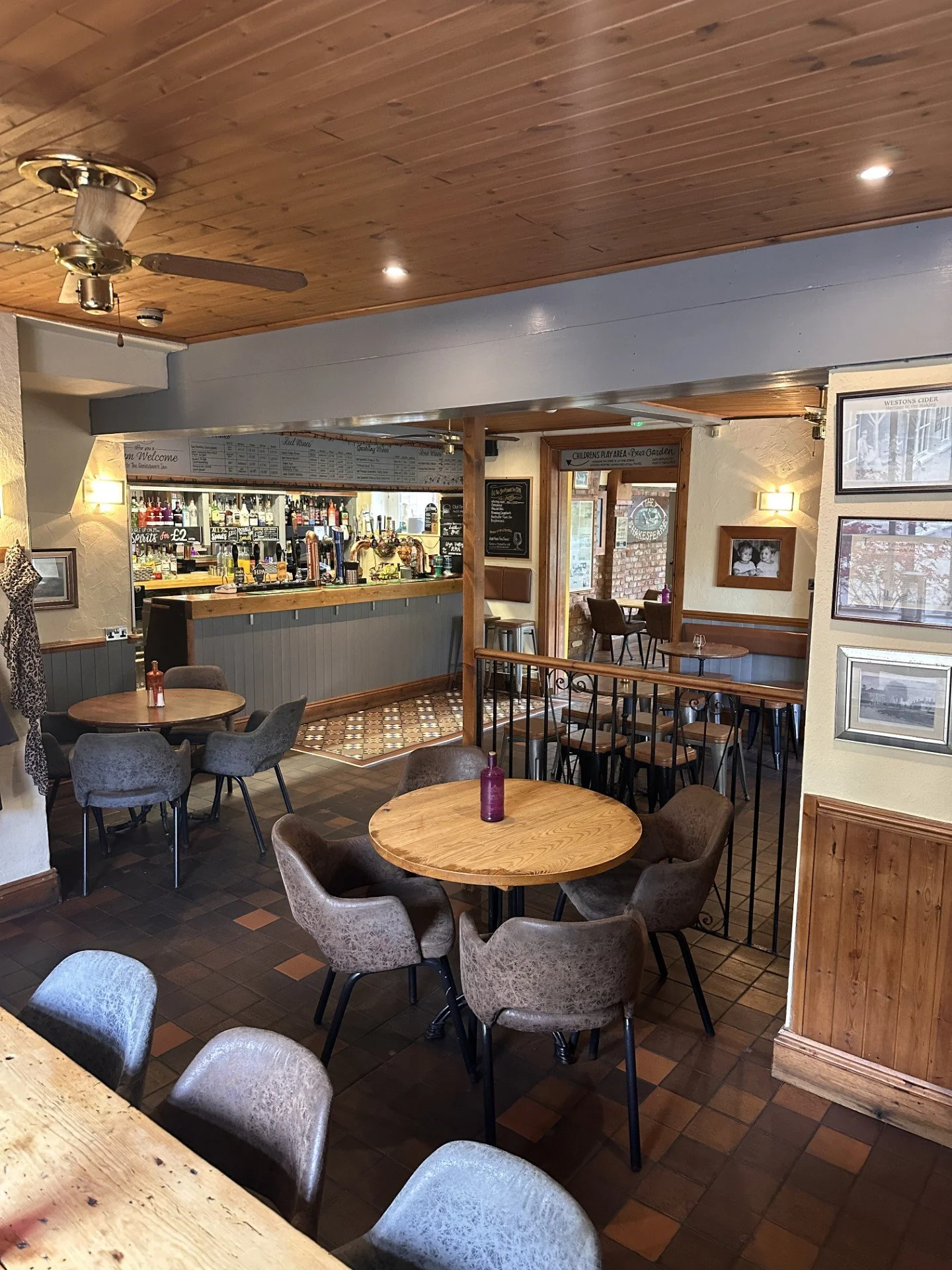 Interior view of a cozy, rustic restaurant or pub with wooden ceiling and furniture, featuring tables with purple bottles, a bar area with various beverages, and framed pictures on the walls.