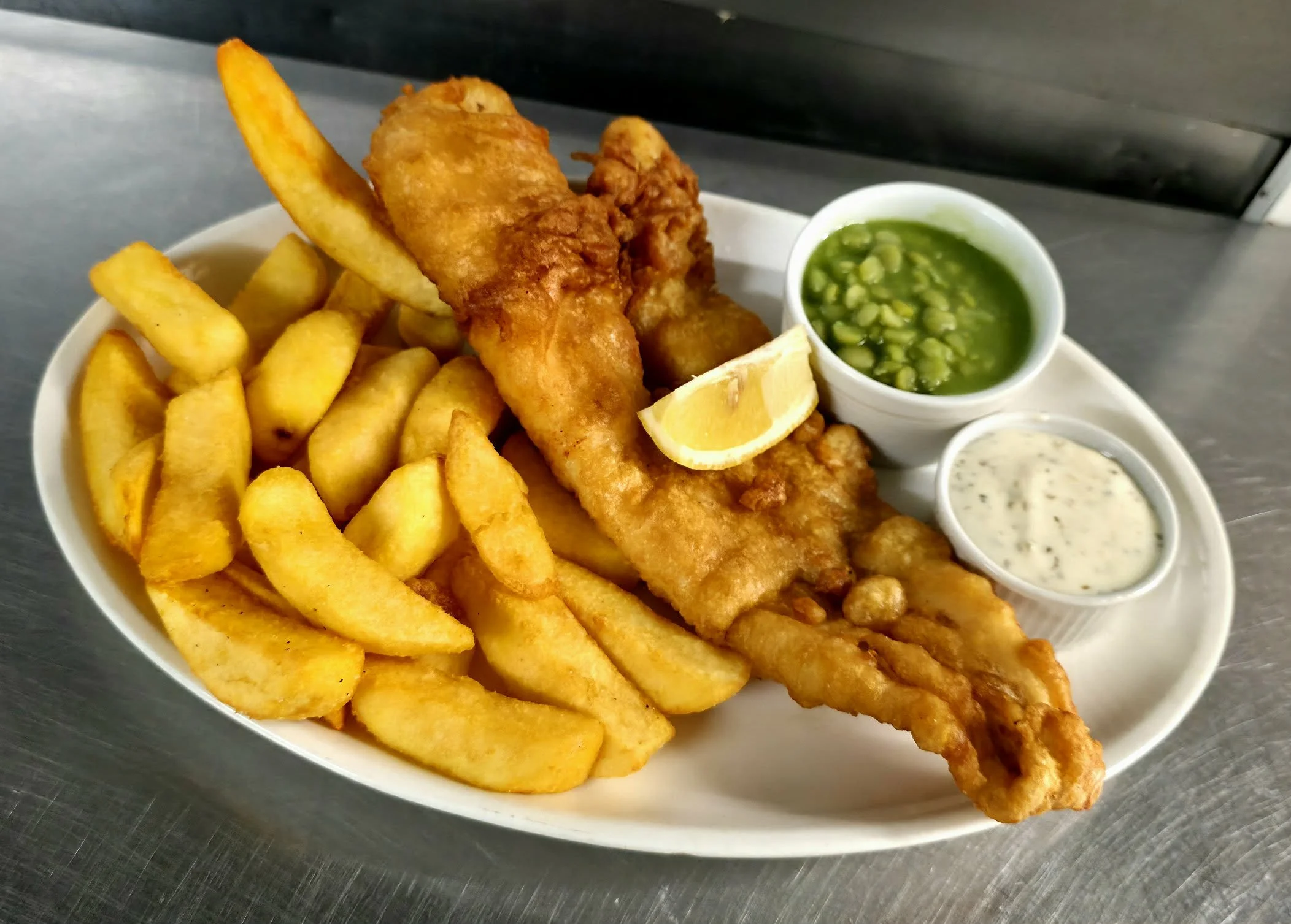 Battered fish fillet, French fries, lemon wedge, green peas in cup, tartar sauce, on white oval plate