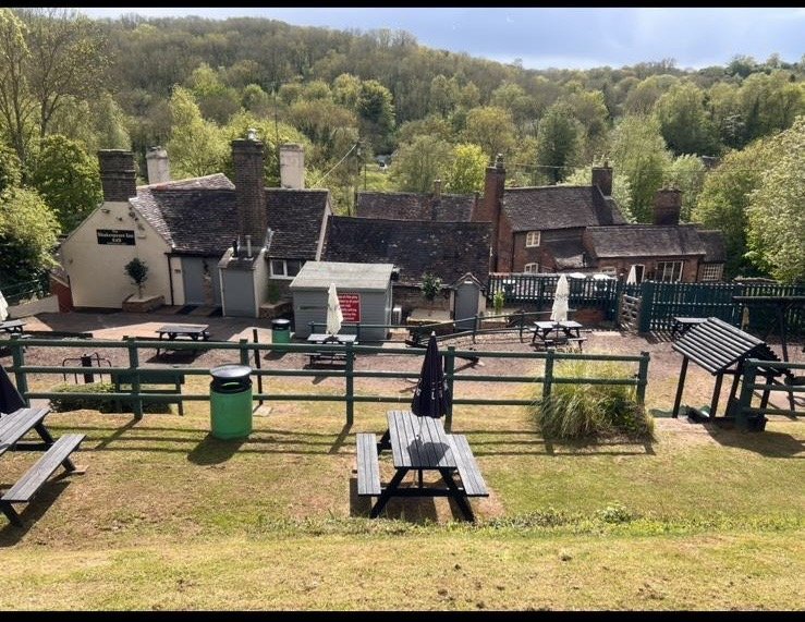 Outdoor seating area with picnic tables, umbrellas, and a fence overlooking a hill with trees in the background.