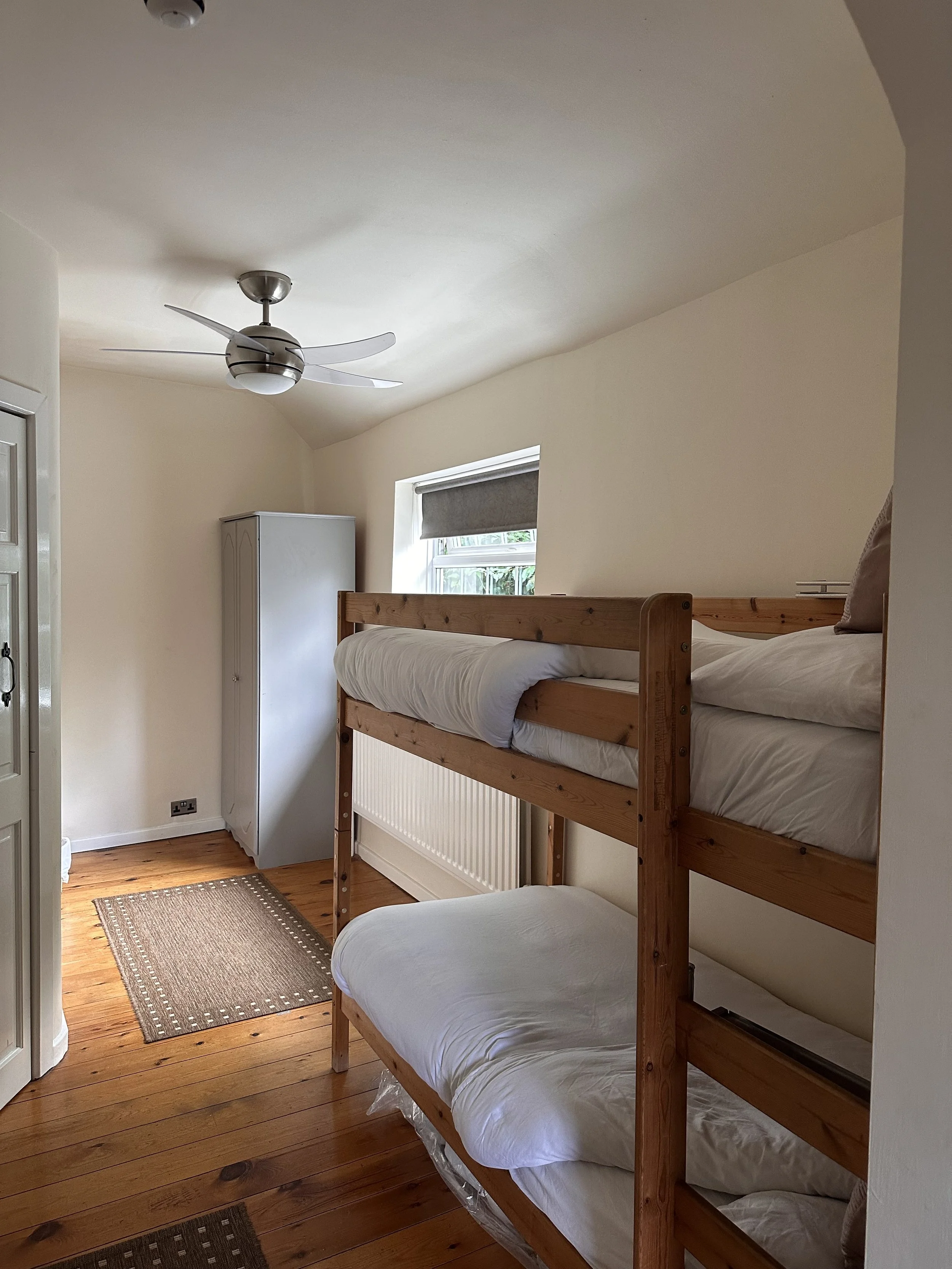 A small bedroom with a wooden bunk bed, a window with a gray roller shade, a white wardrobe, wooden floorboards, and a ceiling fan.