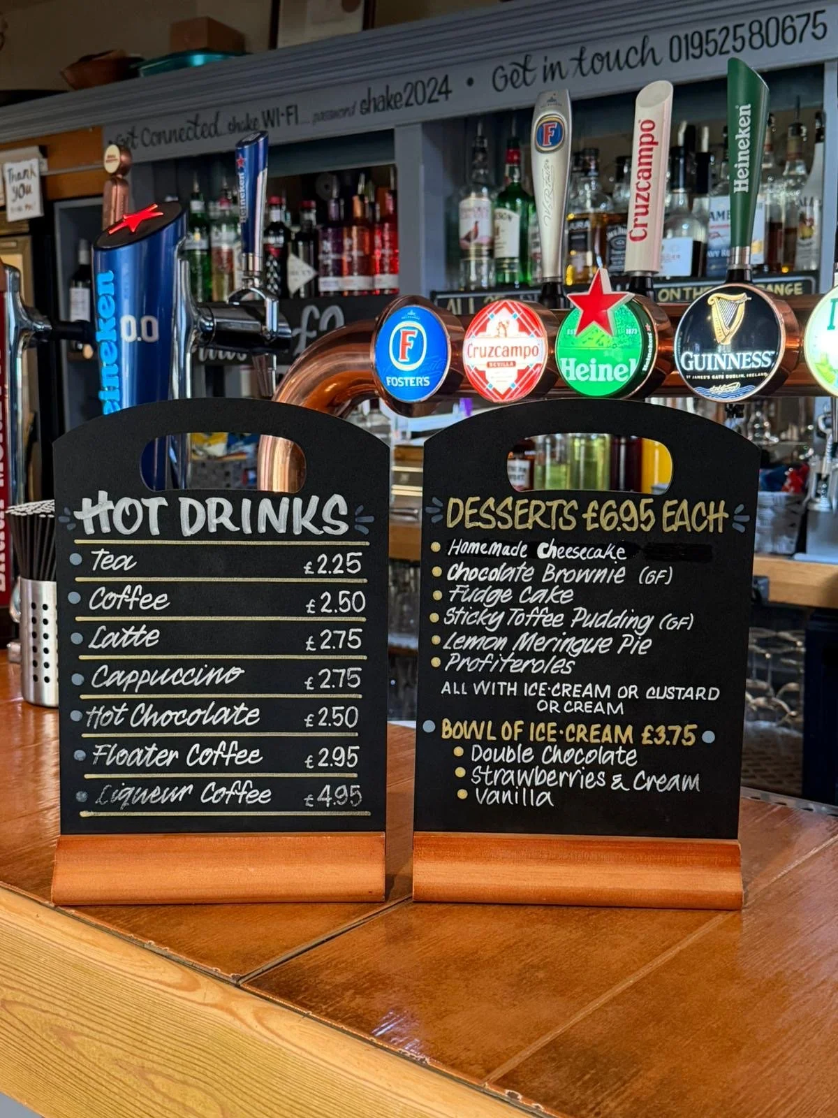 Drink menu boards on a bar counter displaying hot drinks and desserts, with beer tap handles above.