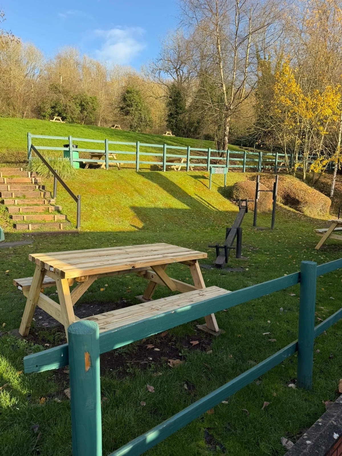Outdoor park area with wooden picnic tables, surrounded by grass and trees, a small hill with stairs, and a sunny clear sky.