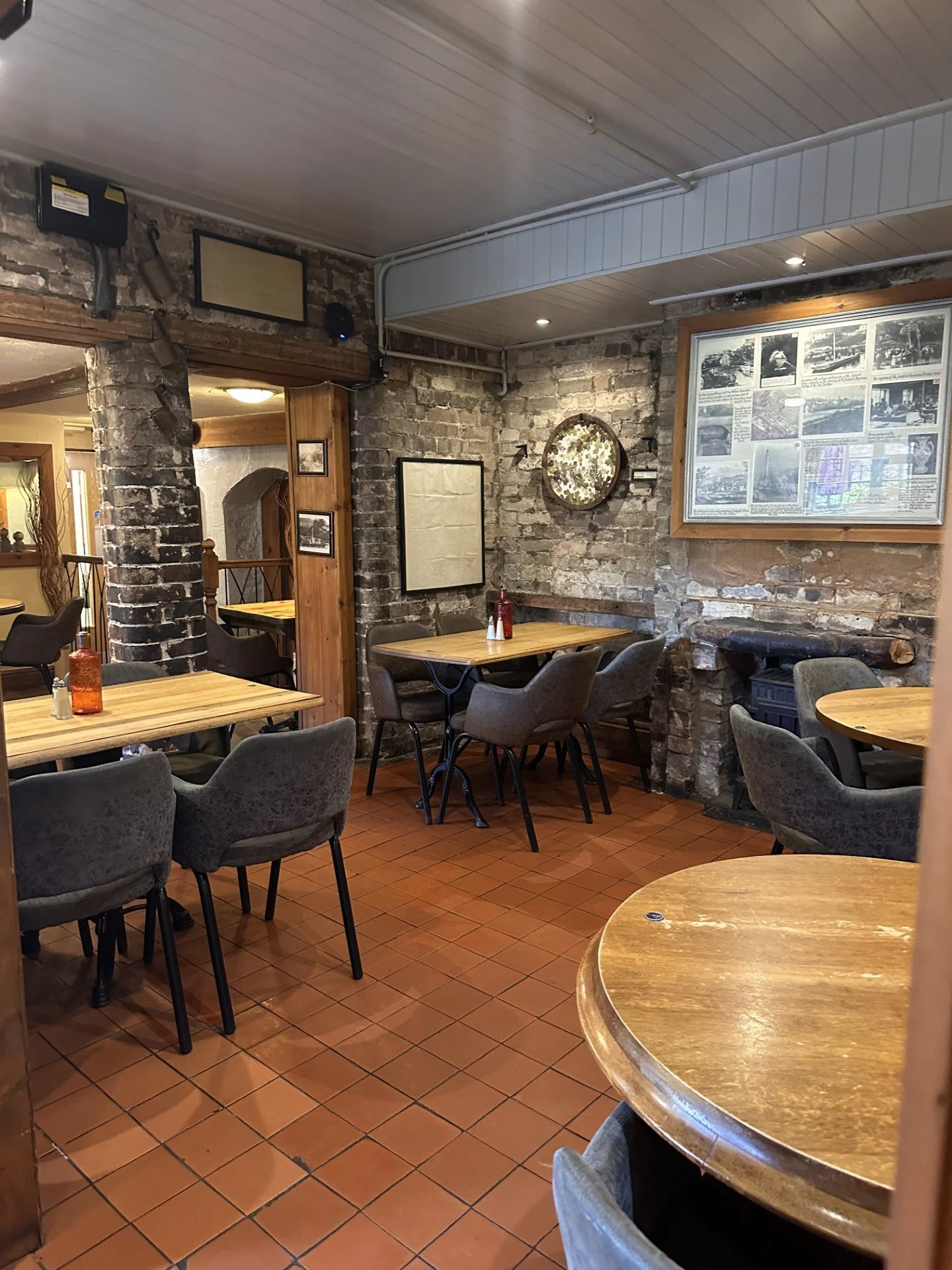 Interior of a cozy restaurant with exposed brick walls, wooden tables with gray upholstered chairs, framed photographs and a large photo collage on the walls, and a terra cotta tiled floor.