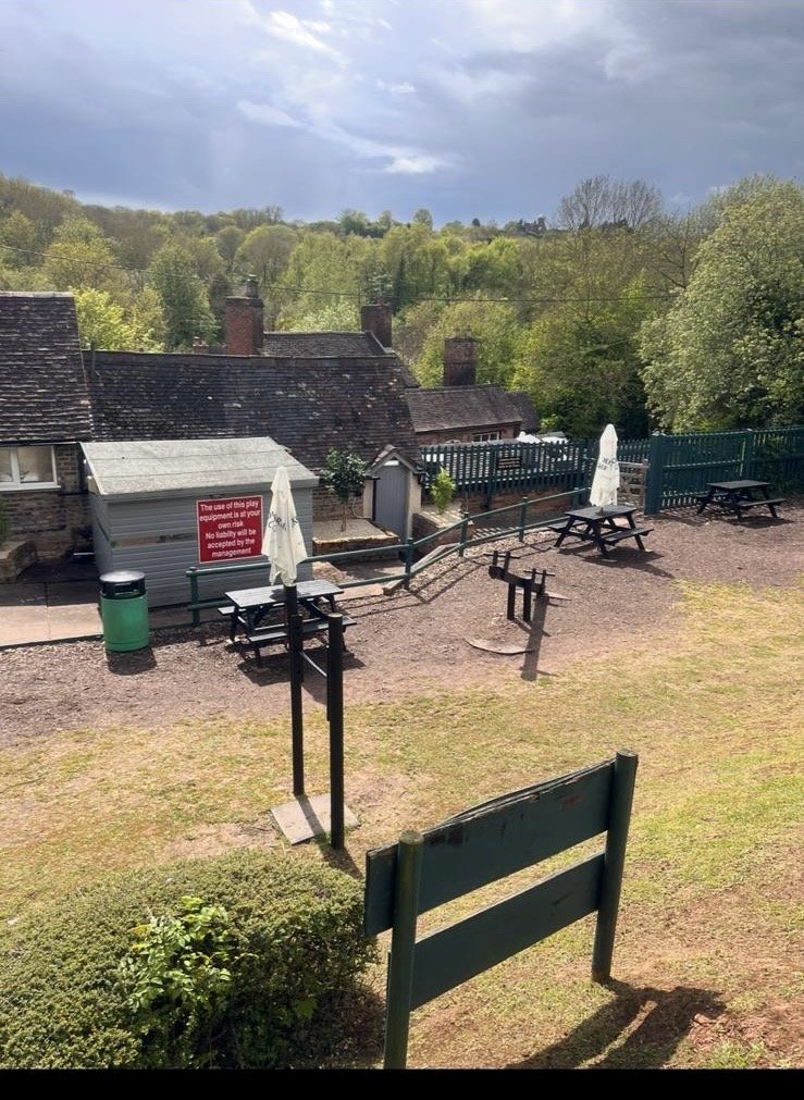 Outdoor area on a hill with several picnic tables, umbrellas, a small playground obstacle course, and a fenced view of rooftops and green trees under a cloudy sky.