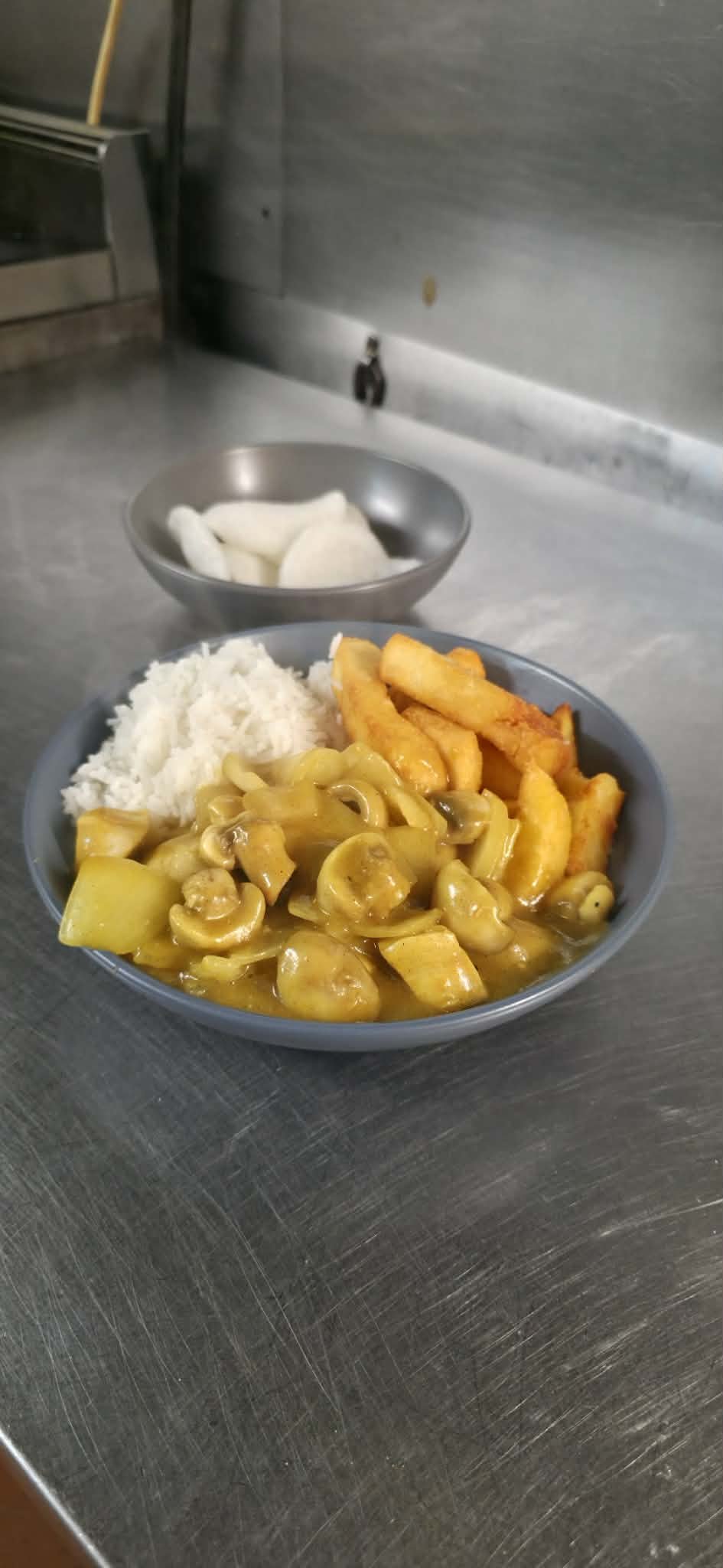 A blue bowl containing rice, fried potato fries, and a mushroom and zucchini stir-fry placed on a stainless steel surface, with another small bowl of sliced onions in the background.