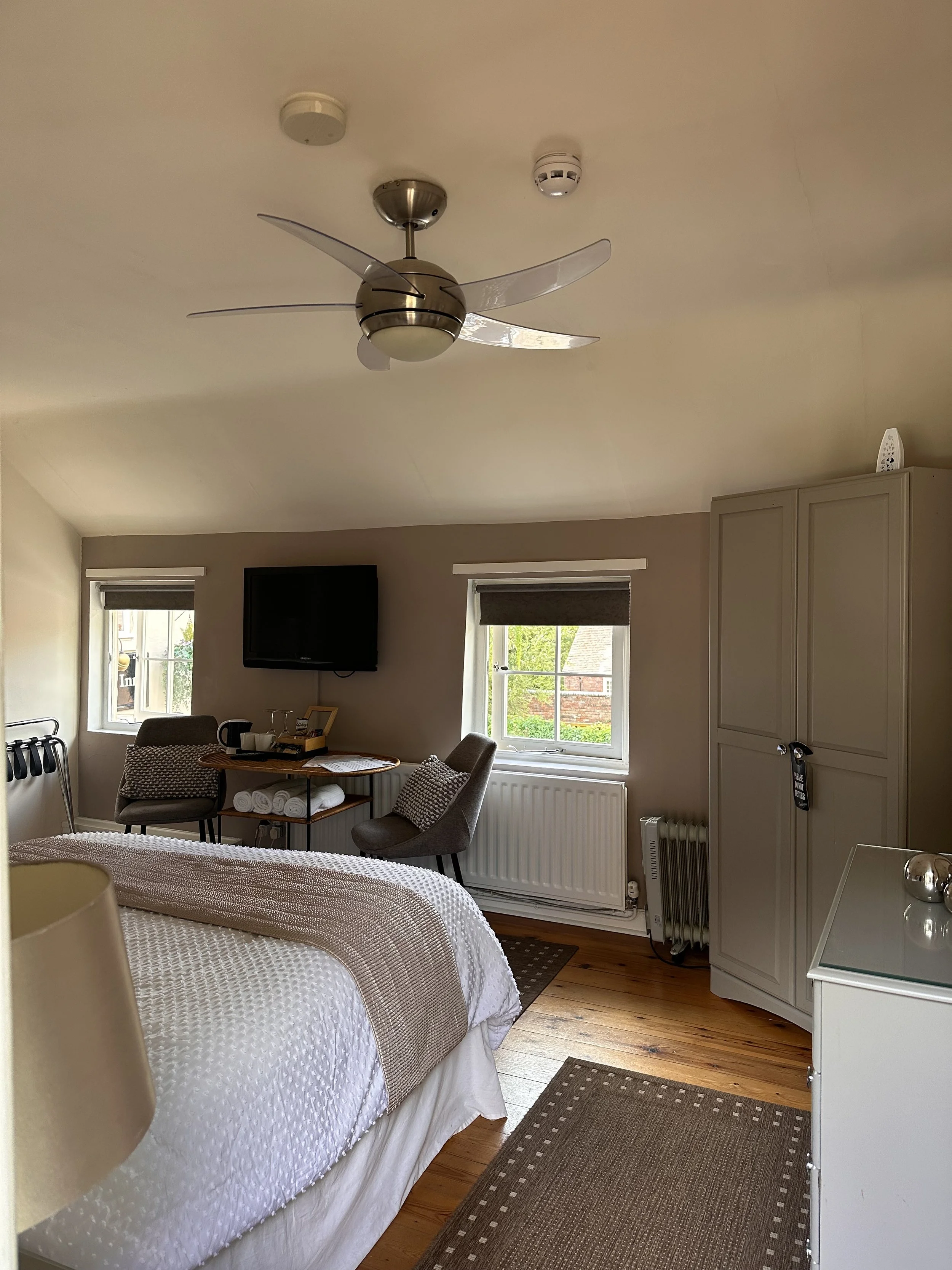 A cozy bedroom with a bed, two windows with brown blinds, a mounted flat-screen TV, a small table with chairs, and a wardrobe. The room has wooden flooring and a ceiling fan with metallic blades.