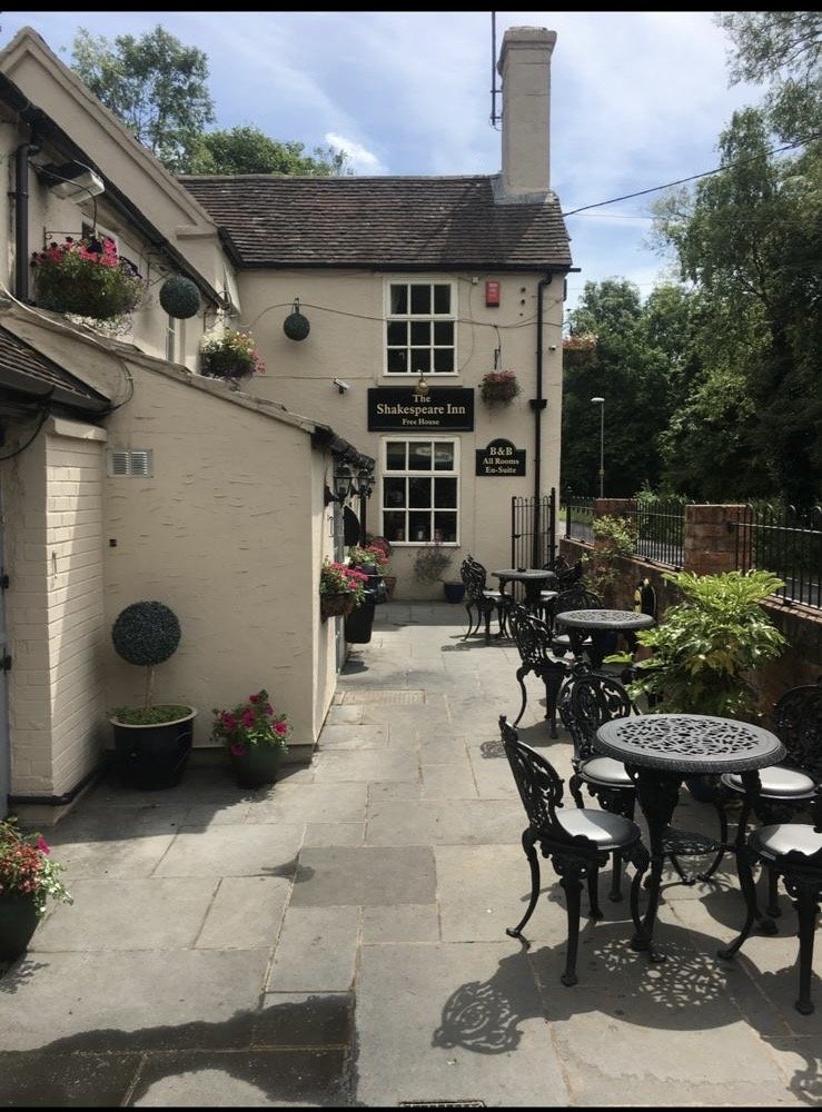 Outdoor patio area of The Shakespeare Inn with black wrought-iron tables and chairs, potted plants, and hanging flower baskets, in front of a white building with multiple windows and a sign for the inn.
