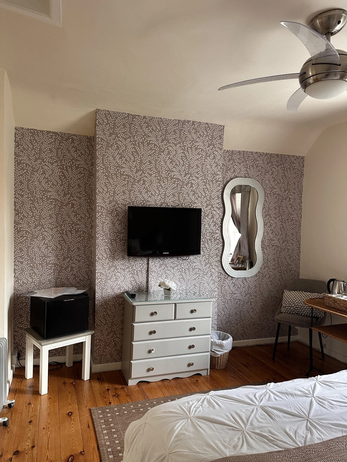 A bedroom corner with a wall-mounted TV, a white dresser, a mirror, a small side table, and a chair. The room has wooden floors and patterned wallpaper.