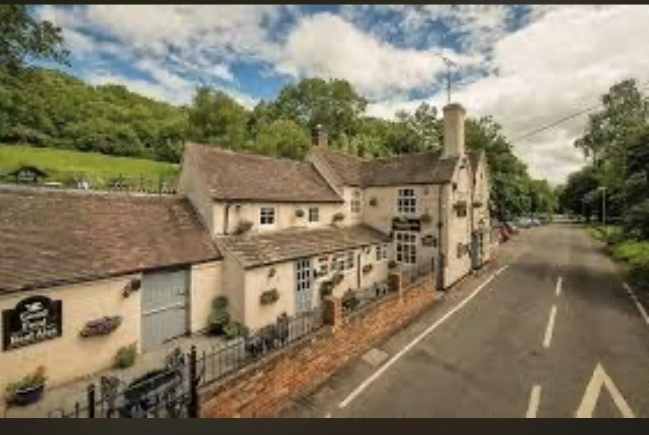 A rustic stone building with a tiled roof near a street, surrounded by green trees and hills under a partly cloudy sky.