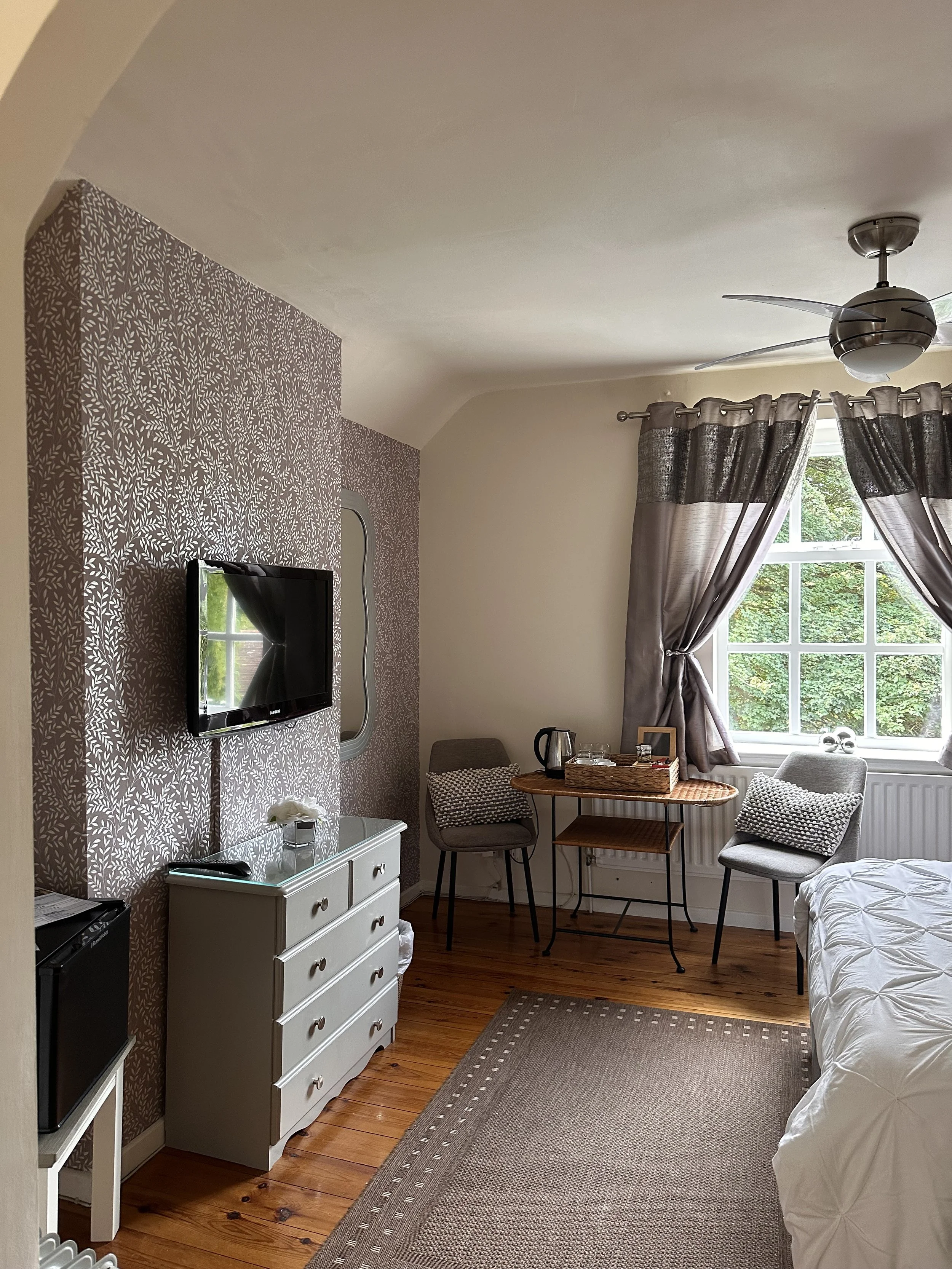 A cozy hotel room with a window, gray curtains, a small table with a water kettle, two chairs, a dresser with a TV, and a bed with a white duvet.
