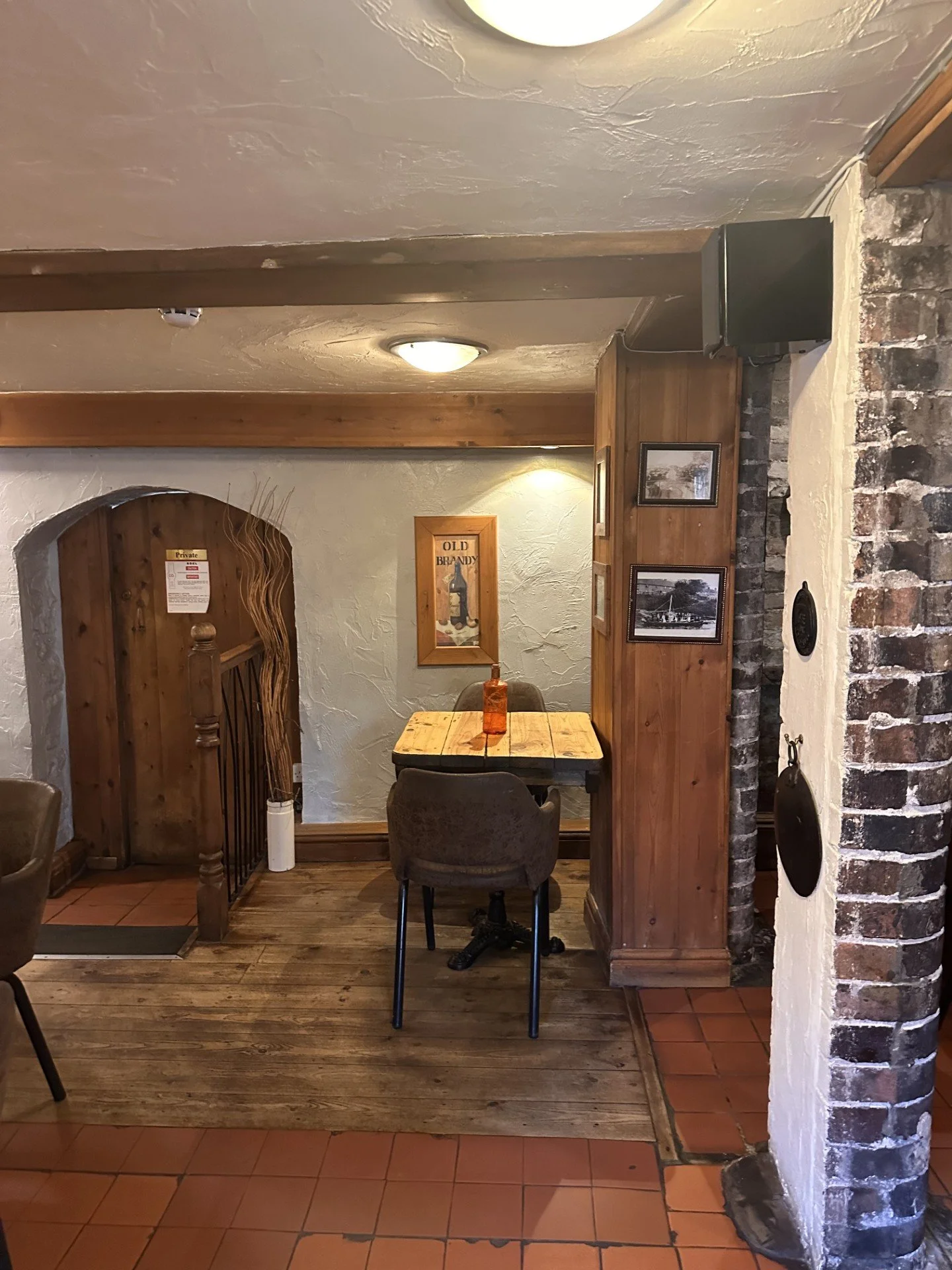 A cozy interior scene of a corner in a restaurant or cafe with wooden and brick decor, a small table with a bottle on it, surrounded by chairs, and framed pictures on the wall.