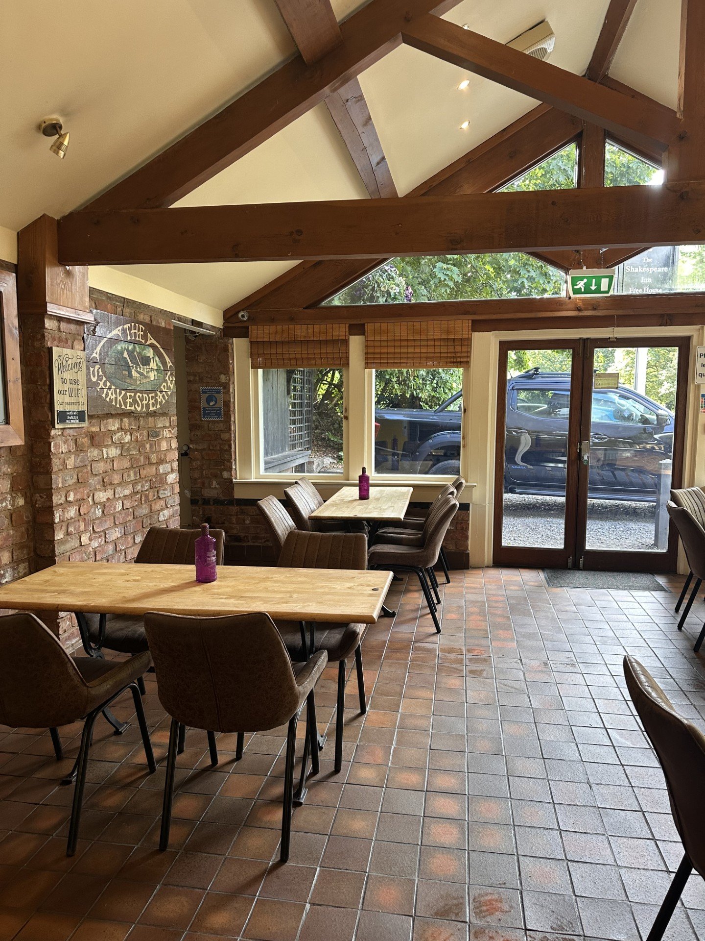 Inside a cozy cafe with wooden beams, brick walls, and large windows. Several tables and chairs are arranged, with a purple water bottle on each table. Outside the glass doors, a parked black truck is visible. Signs about WiFi and emergency exits are