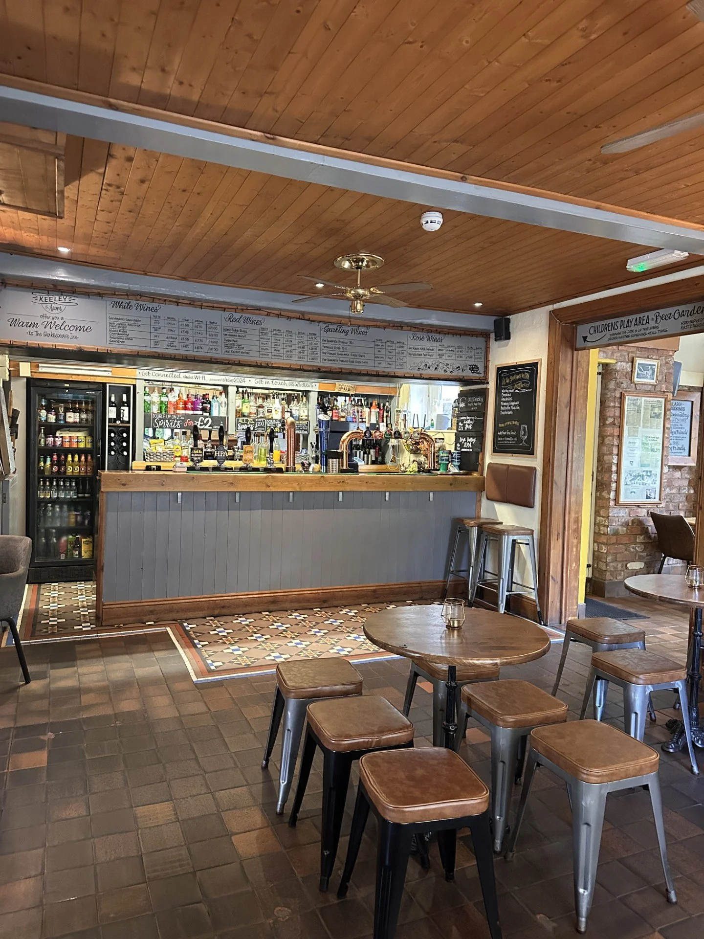 Interior of a cozy pub or cafe with wooden ceiling, bar counter stocked with bottles and taps, tables and chairs, and a brick wall on the side.