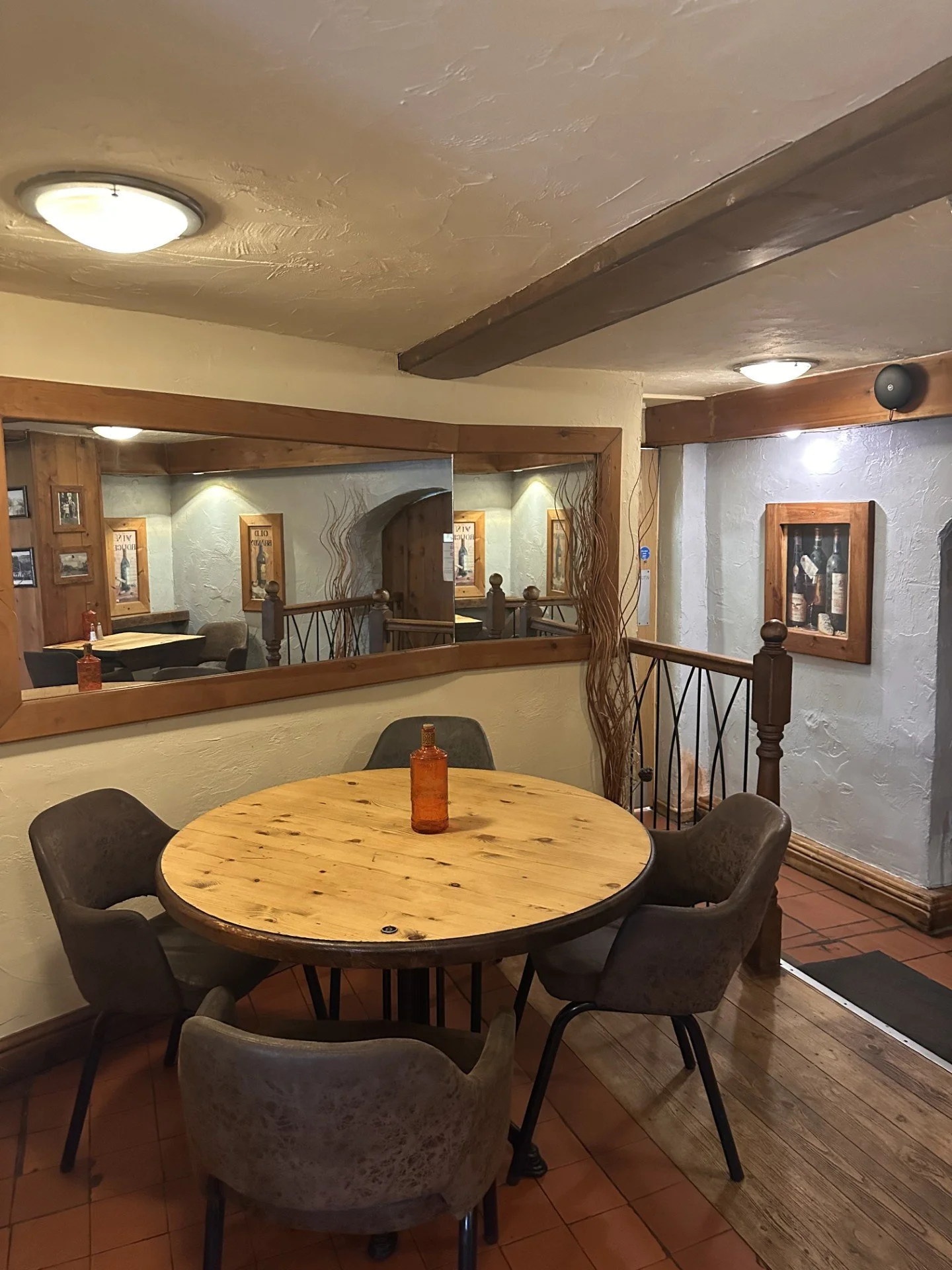 A cozy restaurant dining area with a round wooden table, four upholstered chairs, and a small brown glass bottle in the center. There are wood-framed pictures on the walls, a large mirror, and decorative branches in the corner.