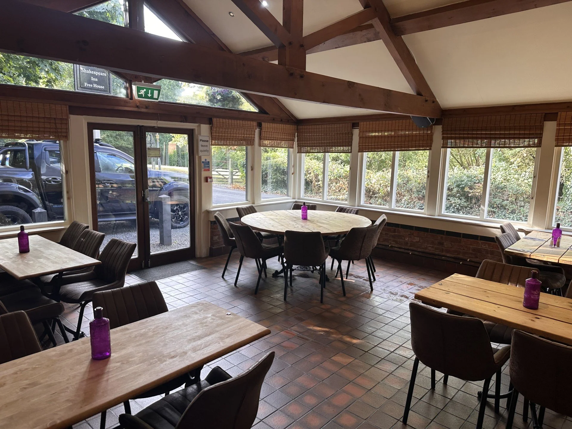 Interior of a cozy restaurant or cafe with wooden tables and chairs, large windows with blinds, and purple water bottles on each table, with a view of greenery outside.