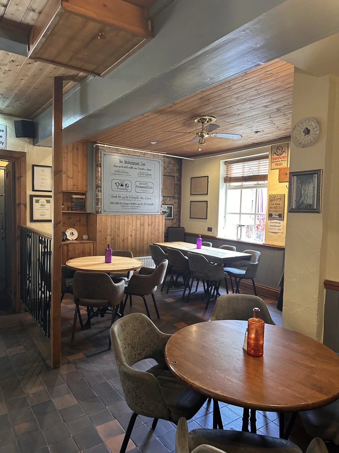 Interior of a cozy café with round and rectangular tables, wooden accents, and a window allowing natural light. There are purple bottles on the tables and various framed items on the walls.