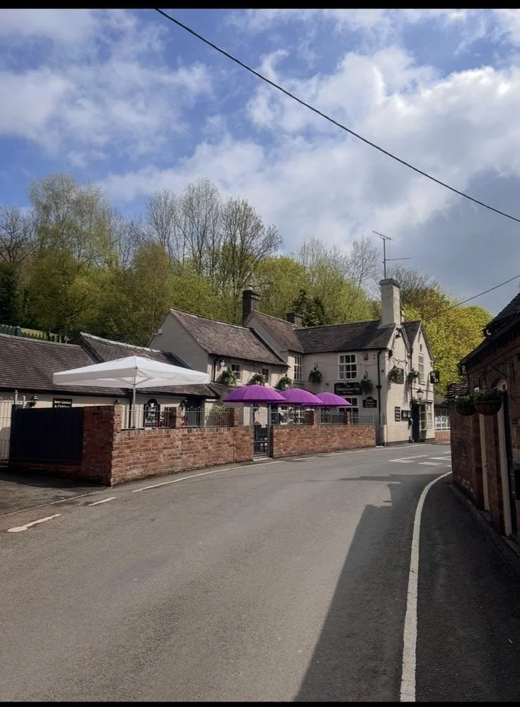A small street with a brick wall and colorful umbrellas. White and purple umbrellas are over outdoor seating, with houses and trees in the background under a partly cloudy sky.
