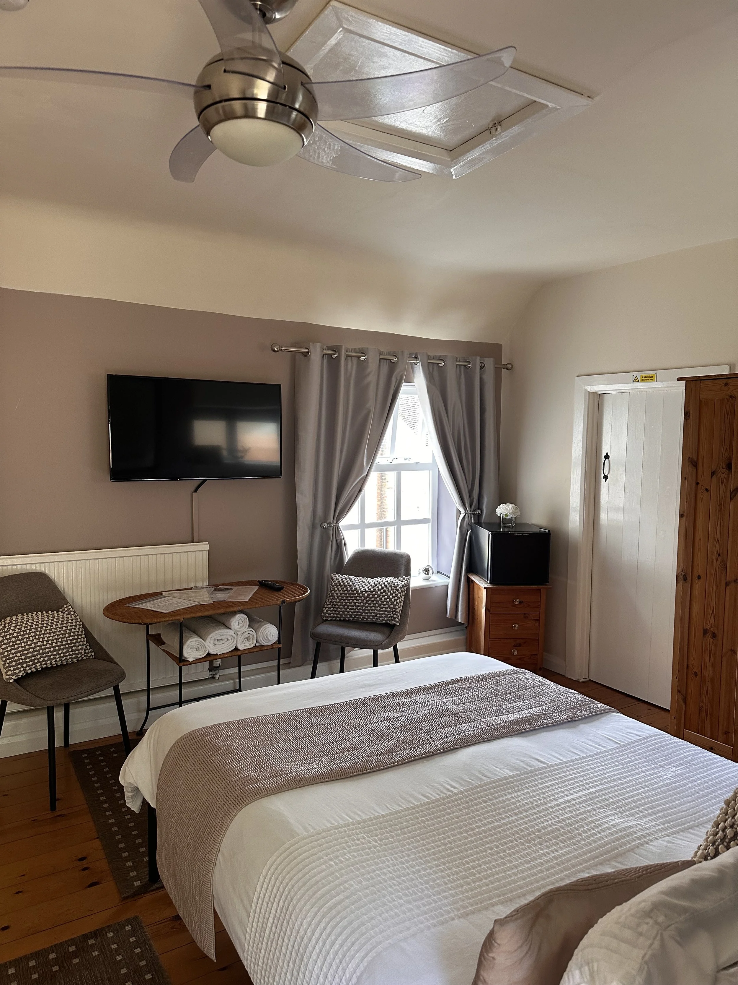 A cozy bedroom with a bed made up with white and beige linens, a nightstand, a wall-mounted flat-screen TV, a window with curtains, two chairs, and a ceiling fan with lights.