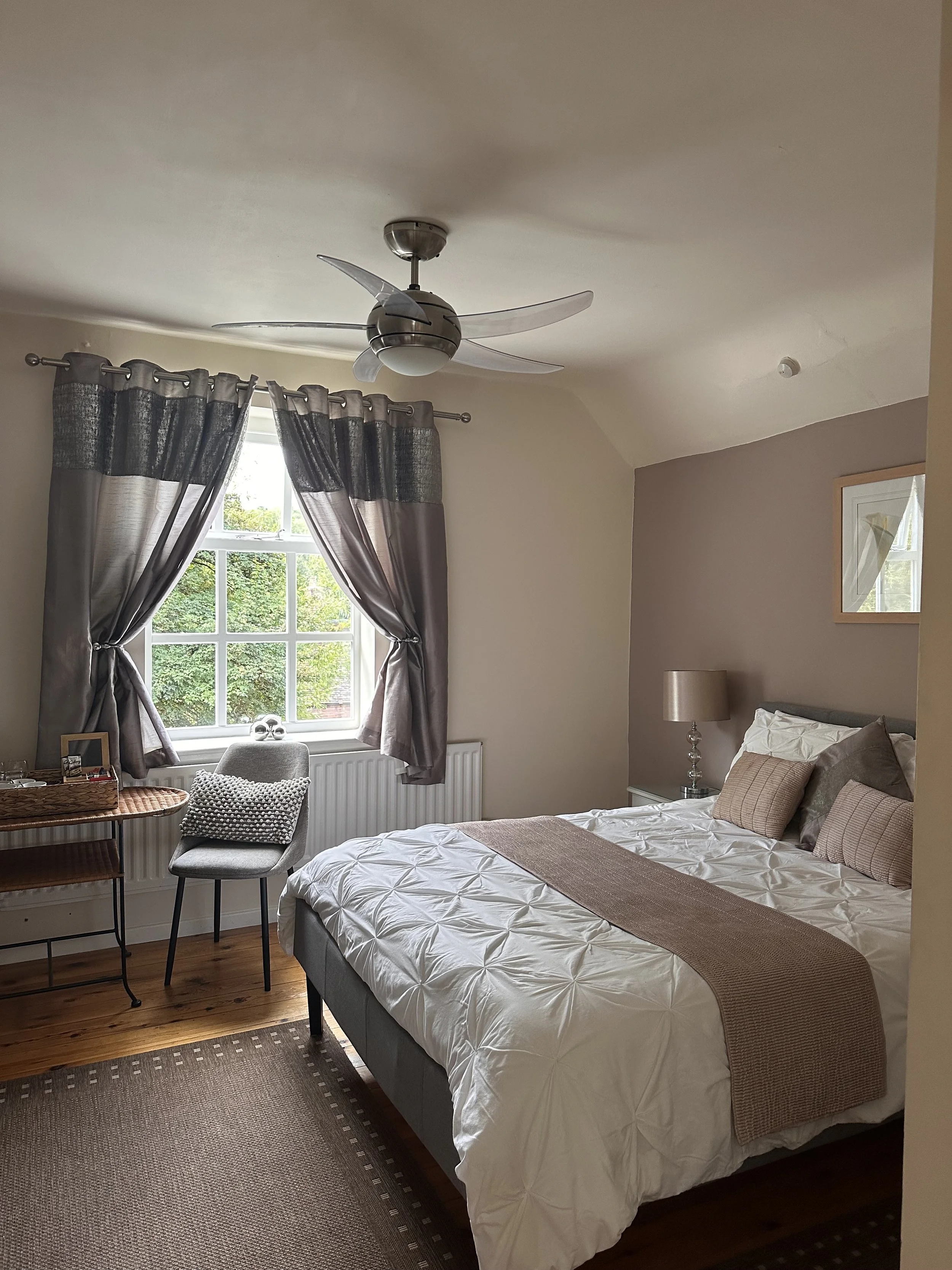 A bedroom with a bed, window with curtains, ceiling fan, chair, and nightstand with lamp.