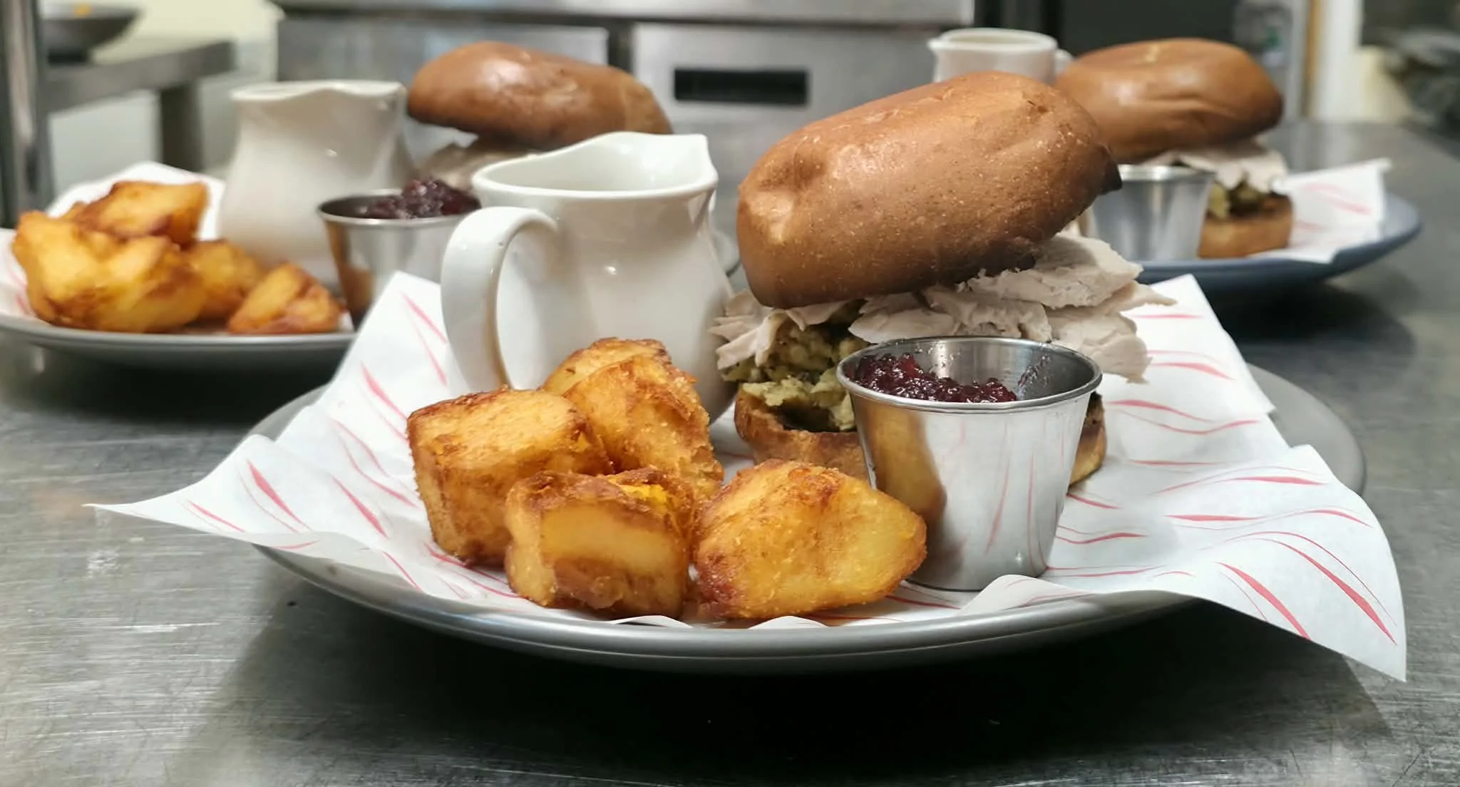 Plate with fried potatoes, turkey sandwich with stuffing in a bun, cranberry sauce, and a small pitcher of gravy or sauce on a metal tray with similar plates in the background.