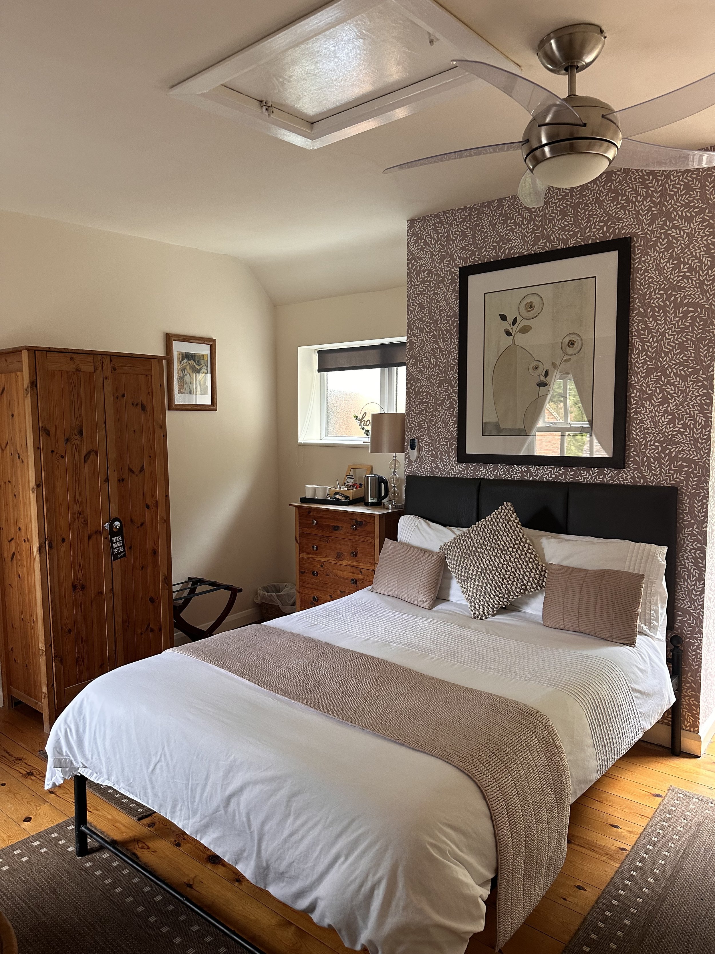 A neatly made bed with a black headboard, white sheets, and four pillows, one patterned and three plain beige, in a cozy bedroom with wooden flooring and furniture. There's a wall art above the bed, a window with a black blind, a wooden dresser with 