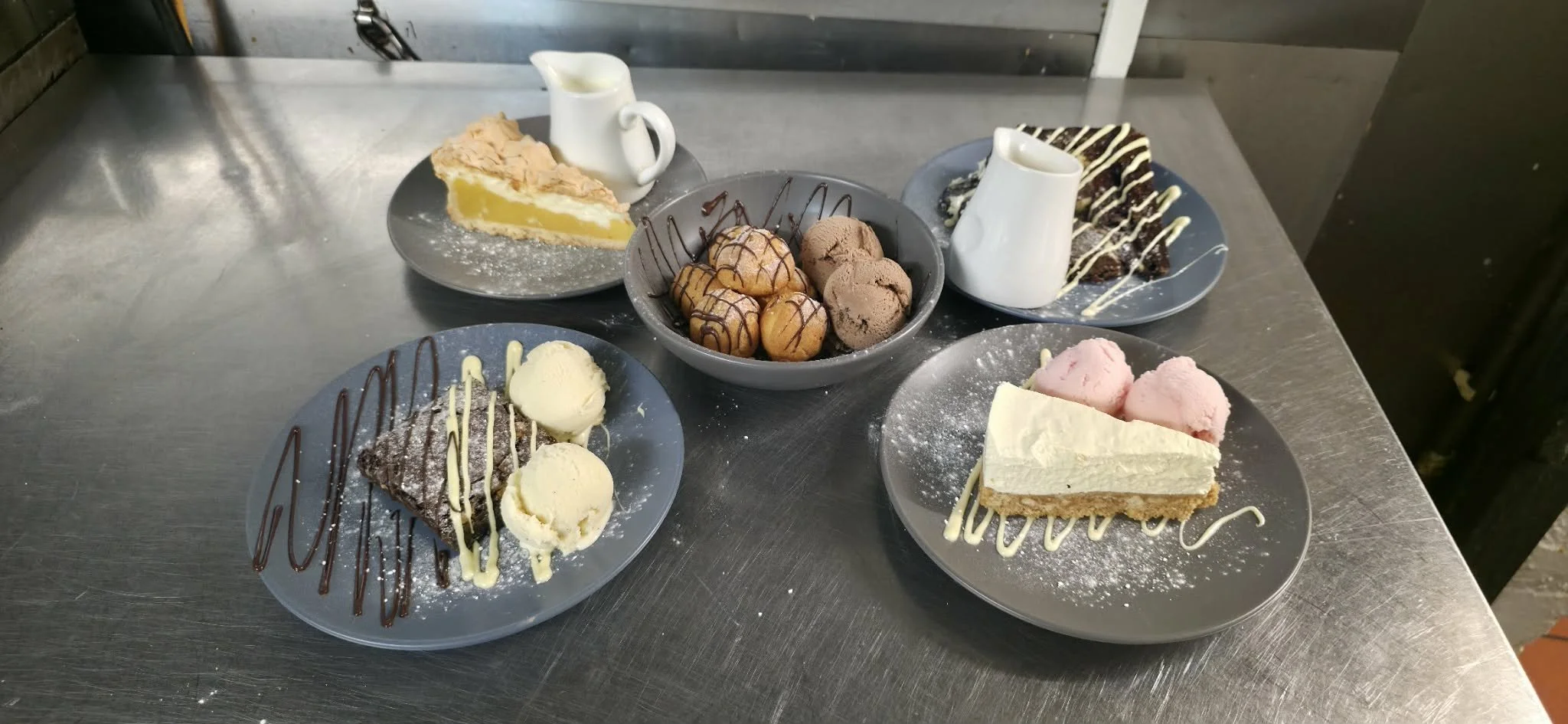 Several plates of assorted desserts on a metal table, including slices of pie, brownies, ice cream, and a bowl of small caramel-drizzled cream puffs.