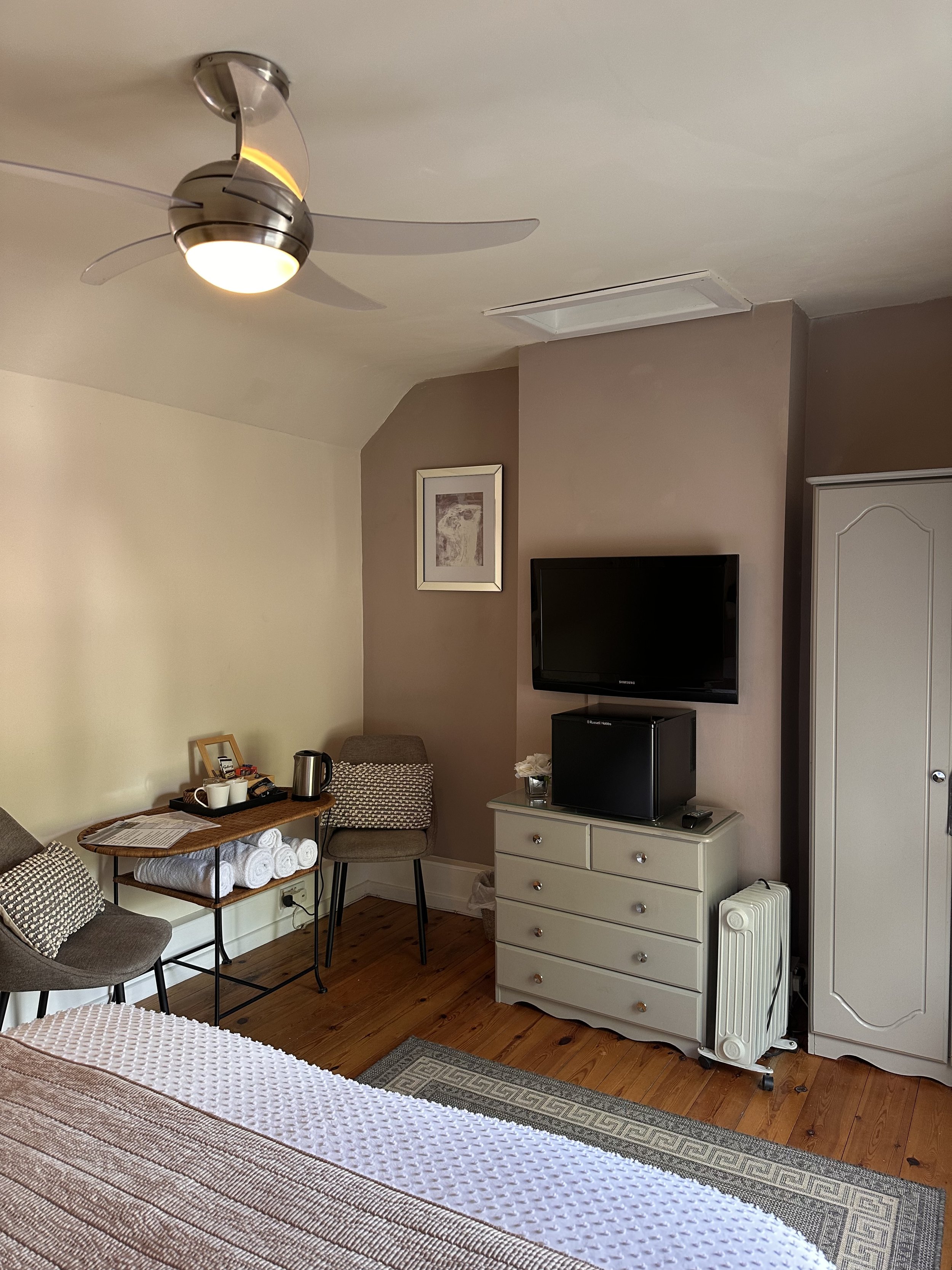 Hotel room with a ceiling fan, a dresser with a mini fridge and TV, a wardrobe, a framed picture, a small table with bathroom amenities, towels, and a chair, part of a bed with a striped blanket, and a hardwood floor.
