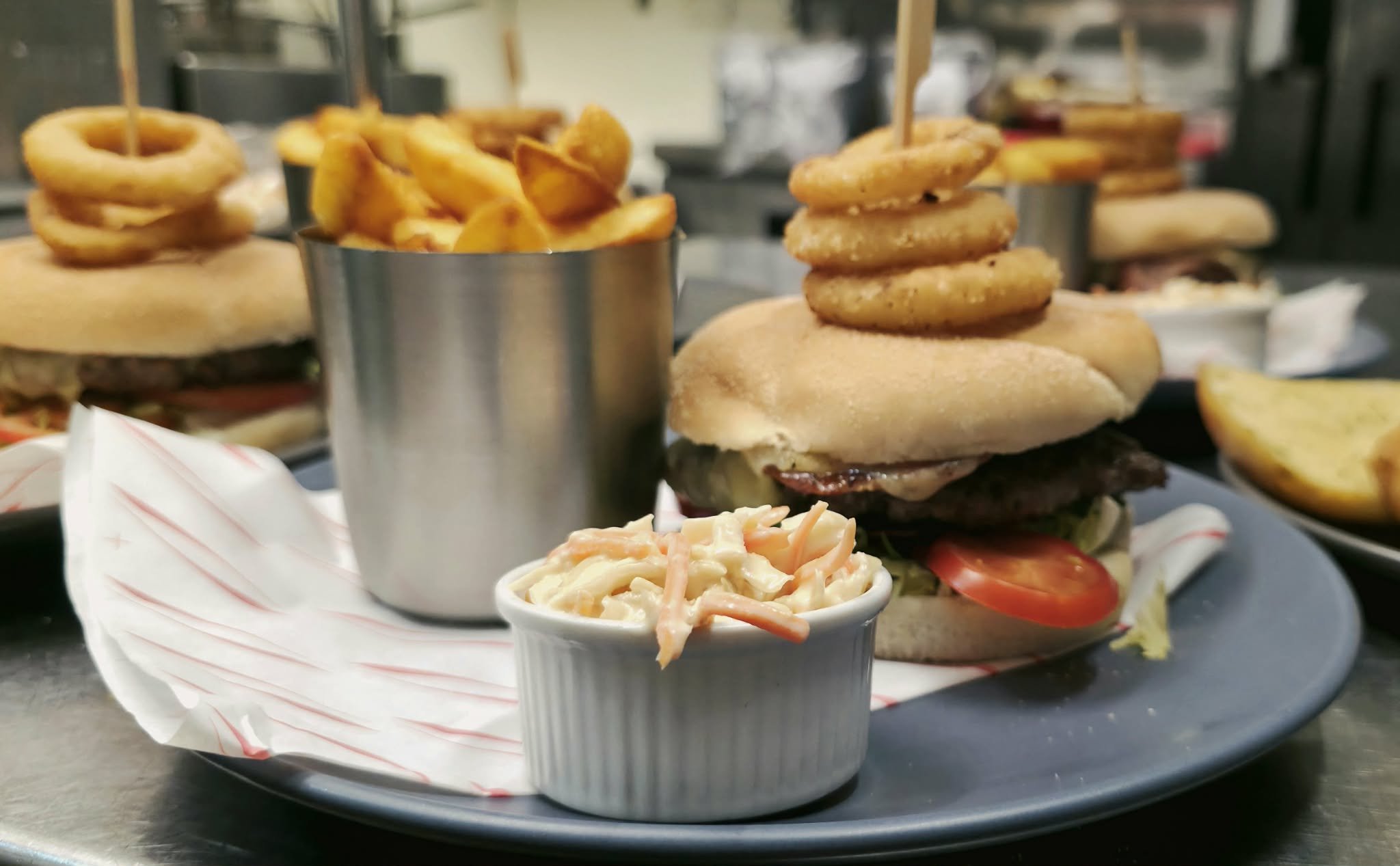 Close-up of a cheeseburger with onion rings stacked on top, served with a side of macaroni salad in a small white cup and a metal cup of French fries on a dark plate.