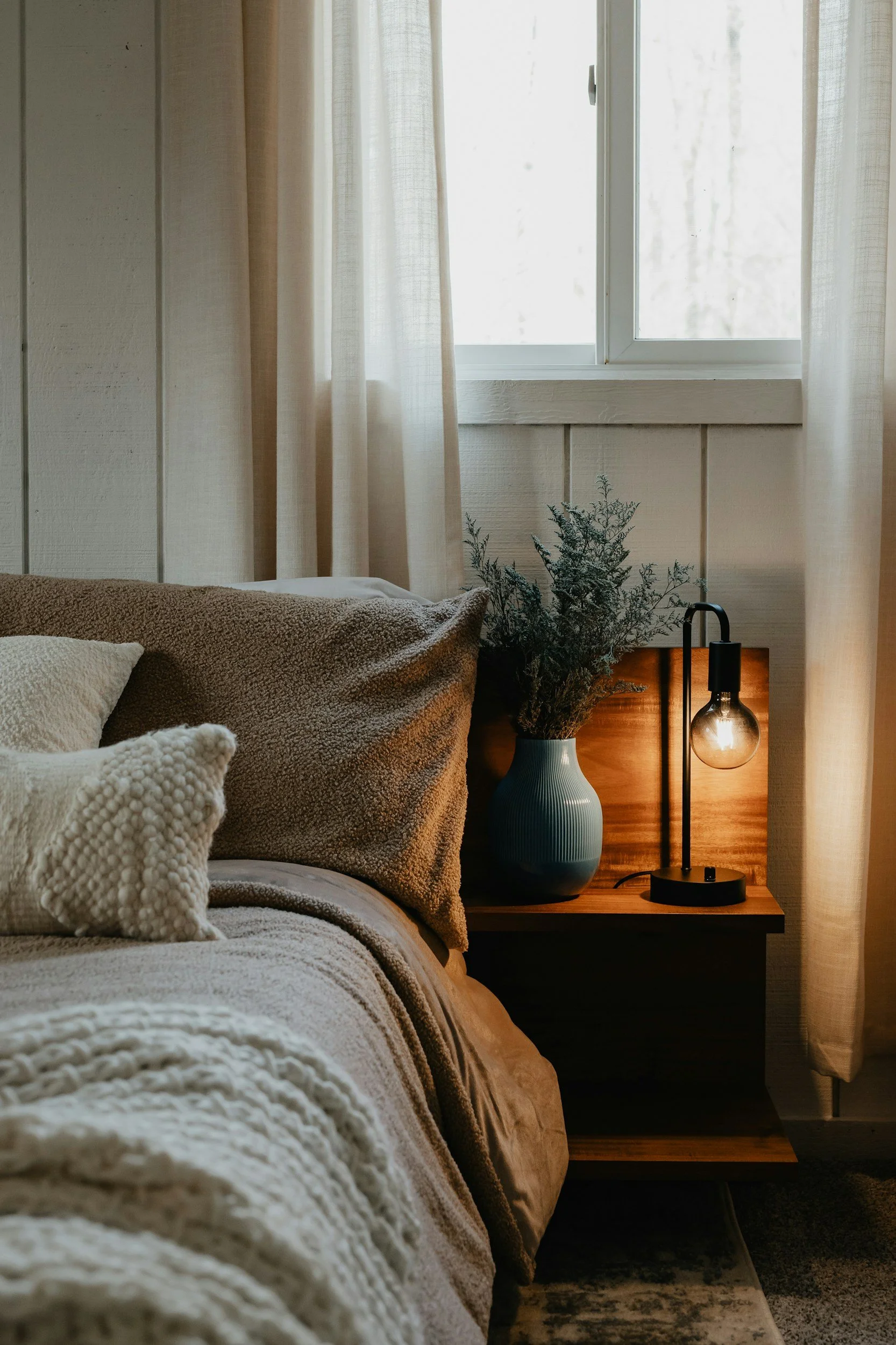 A cozy bedroom corner with a bed covered in neutral-toned blankets and pillows, a wooden side table with a vase of greenery, and a modern lamp with an exposed bulb beside a window with cream curtains.