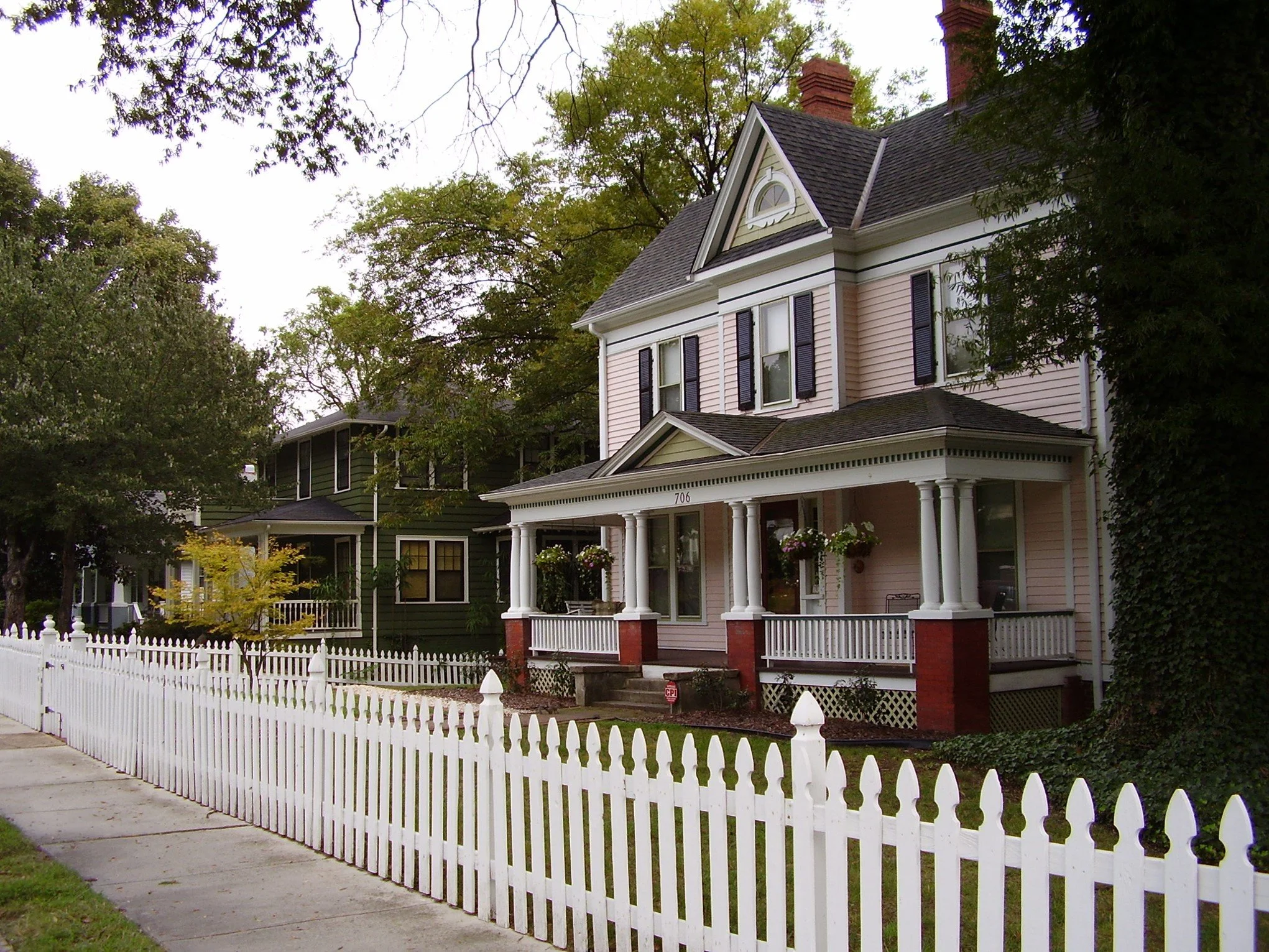 A pink Victorian style house with a porch, white picket fence, and black shutters, surrounded by trees and a sidewalk.