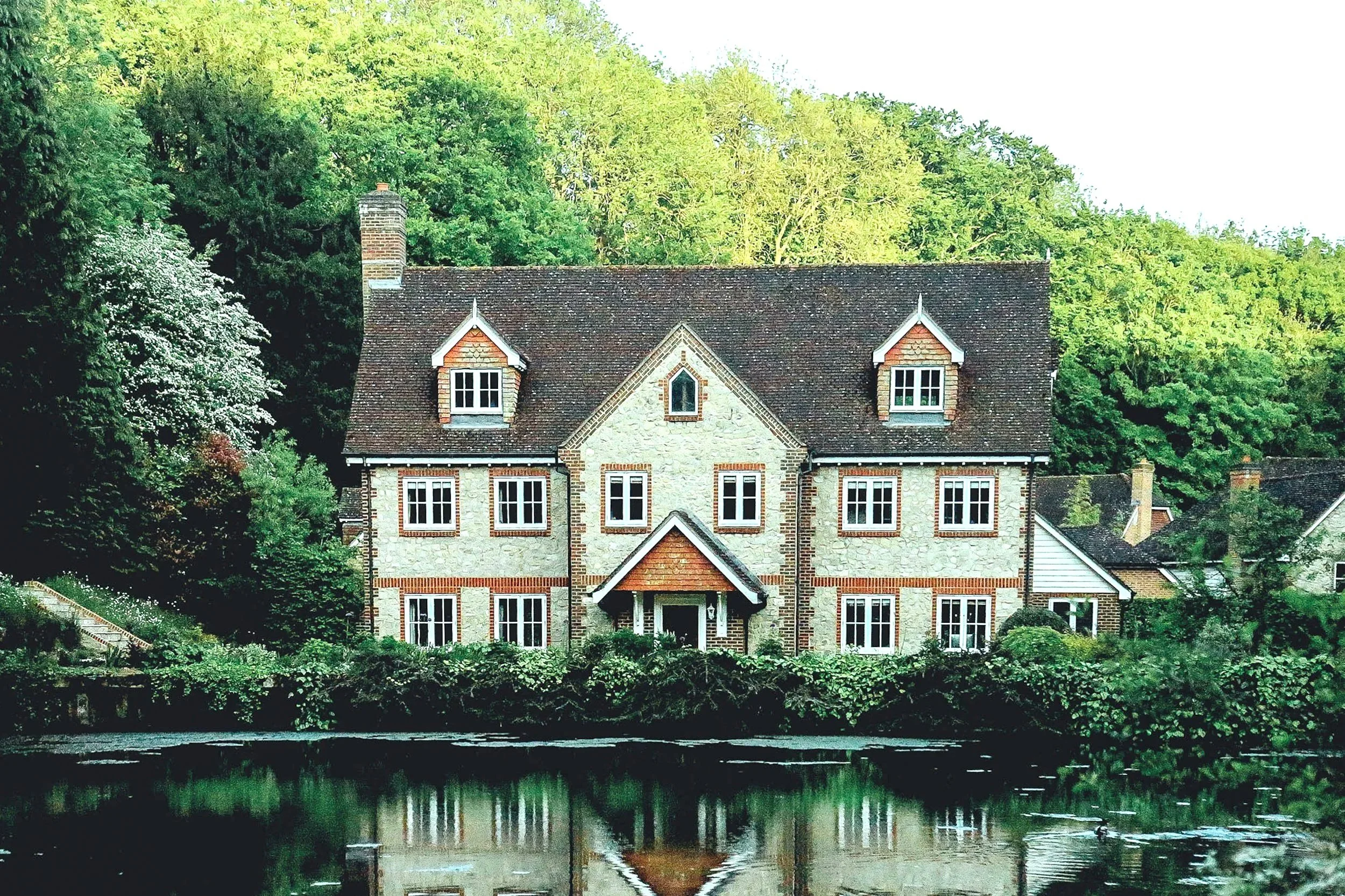 A large historic house with stone and brick exterior, multiple windows, and a steep slate roof, situated near a pond with surrounding greenery and trees.