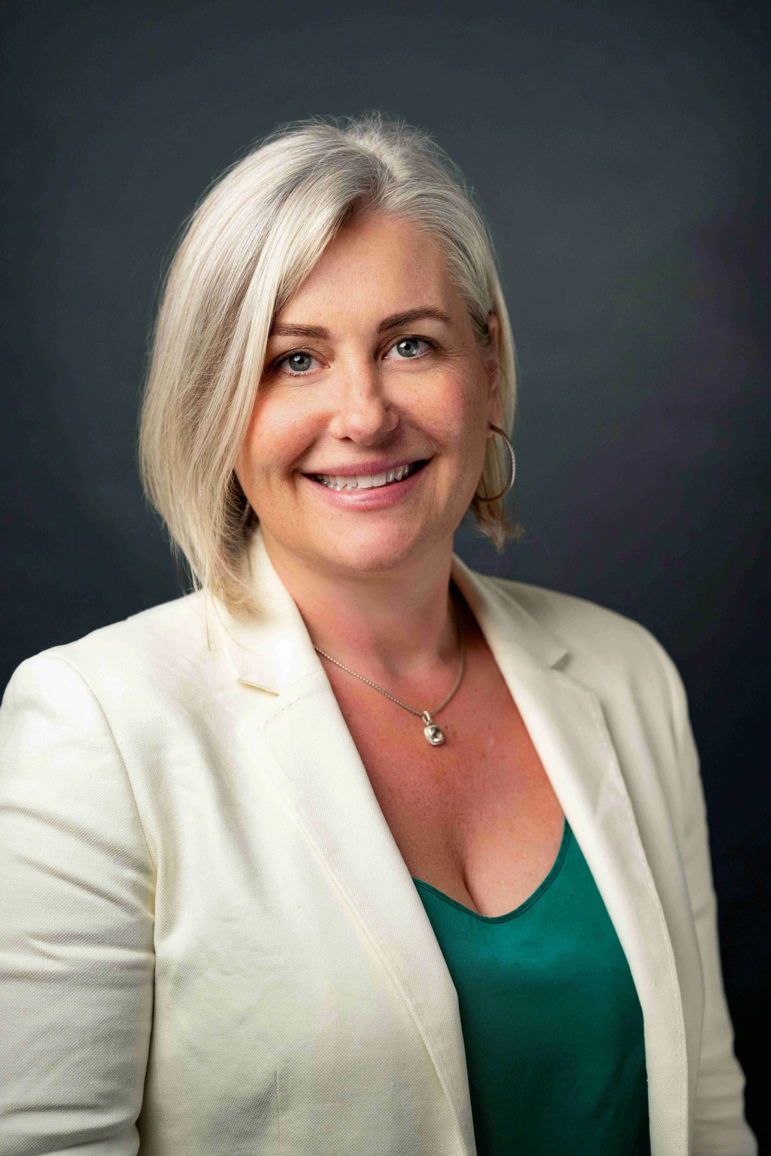 A woman with shoulder-length blonde hair, wearing a cream blazer over a teal top, smiling and posing against a dark background.